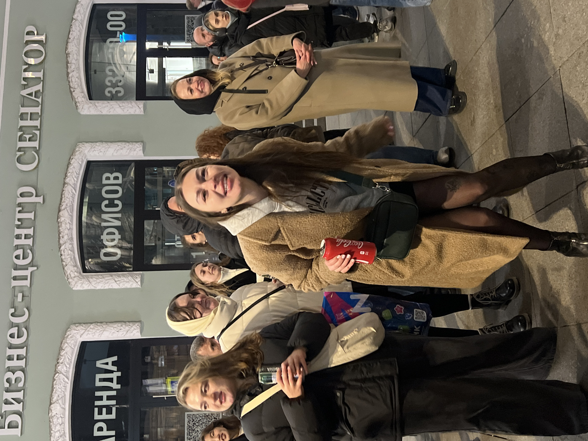 A group of approximately 10 people stand outside of the "Business Center Senator" building in Saint Petersburg, Russia. The architecture indicates a typical business building with windows labeled "Offices" and "Rent." Several women are visible, wearing winter coats in beige, white, and black, suggesting a cool climate. One of the women is holding a red Coca-Cola can. The crowd appears to be diverse, with both men and women of different ages, indicating a casual gathering or waiting for something. The setting is an urban environment, with a tiled sidewalk. The photo appears to be taken in the evening, based on lighting conditions.