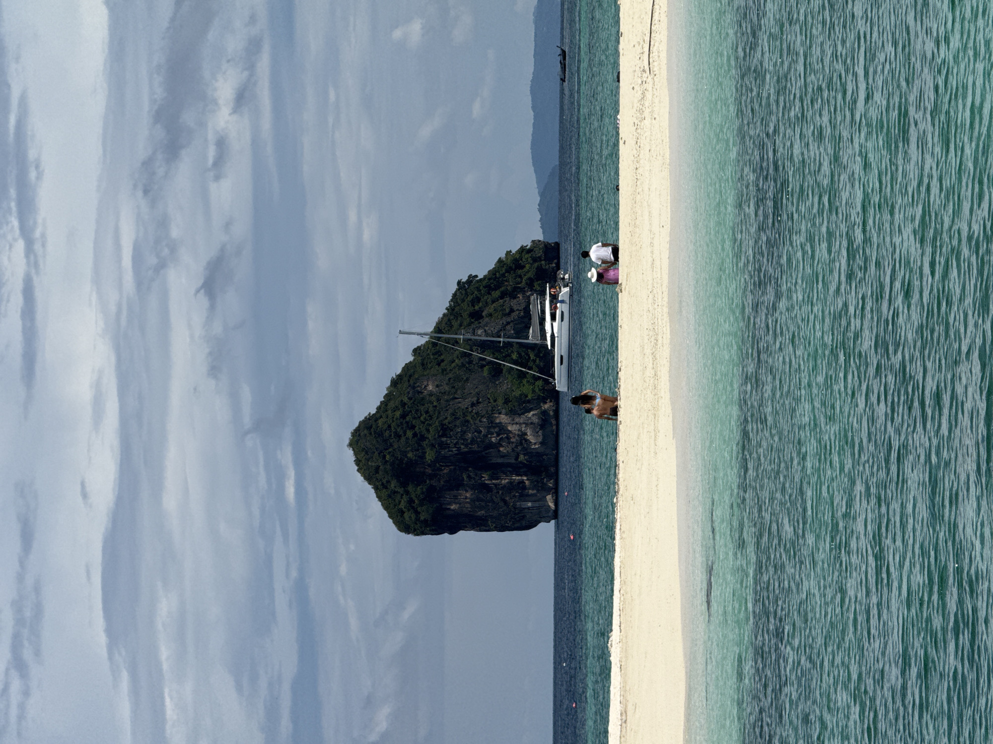 A daytime, medium shot captures a scenic view of a small, lush green island in the sea, with a catamaran anchored nearby, off the coast of Ao Nang, Thailand. The island, with its steep, rocky sides covered in dense vegetation, stands prominently in the center-left of the frame, dwarfed by a cloudy, overcast sky. In the foreground, a strip of white sand beach is visible, where three individuals are seated, facing the water. To the left, a woman in a bikini is seen from behind. Further right, a couple, a man in a white t-shirt and a woman in a pink dress and white hat, sit side-by-side. The water transitions from a shallow, turquoise hue near the shore to a deeper blue further out. Other distant islands and a traditional longtail boat are faintly visible on the horizon.
