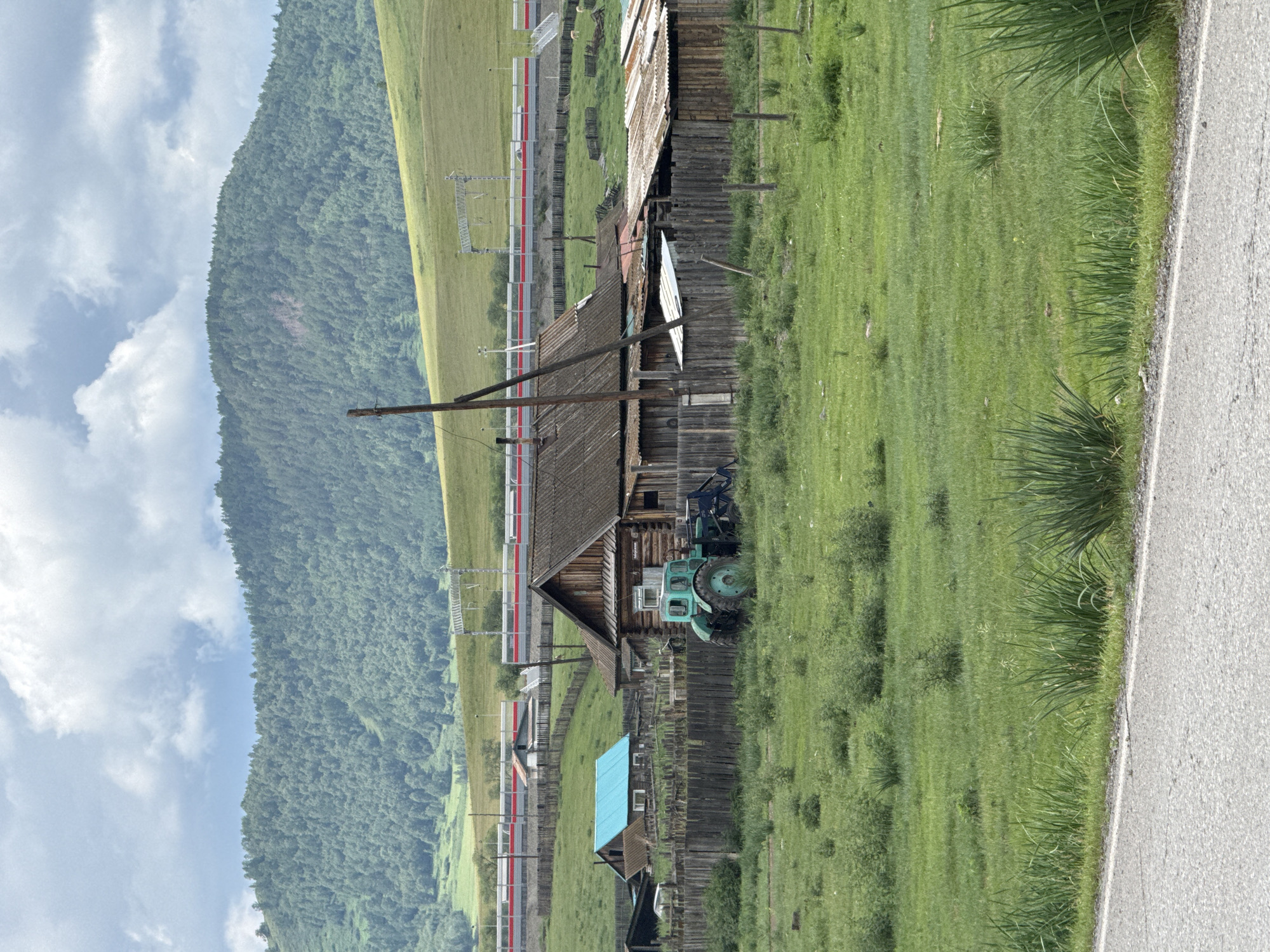 The image depicts a rural village scene in Kazanovka, Russia, under a cloudy sky. In the foreground, a paved road runs horizontally, bordering a grassy field. The field contains scattered green bushes and tall grasses. Further back, a wooden fence encloses a residential area. Within the enclosure, a rustic wooden house with a weathered roof stands prominently. Adjacent to the house is a green tractor with a front loader. Another smaller building with a bright blue roof is visible to the left, also enclosed by the wooden fence. In the mid-ground, a train with red and white stripes travels on elevated tracks. The background is dominated by a lush, green, tree-covered mountain slope.