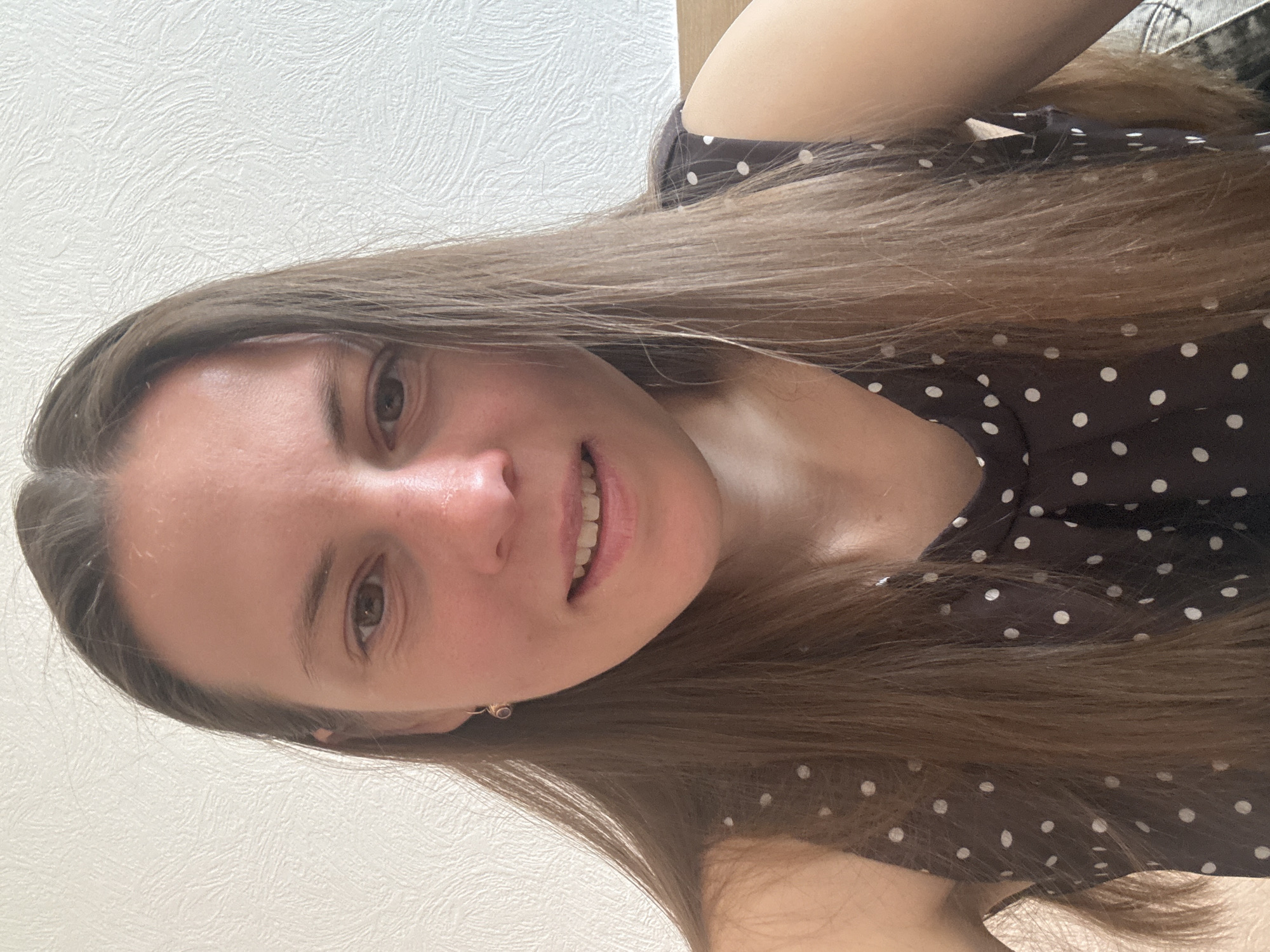 A woman with long, light brown hair and fair skin is smiling and looking directly at the camera. She is wearing a brown polka-dot dress. The background is a textured, off-white wall.
