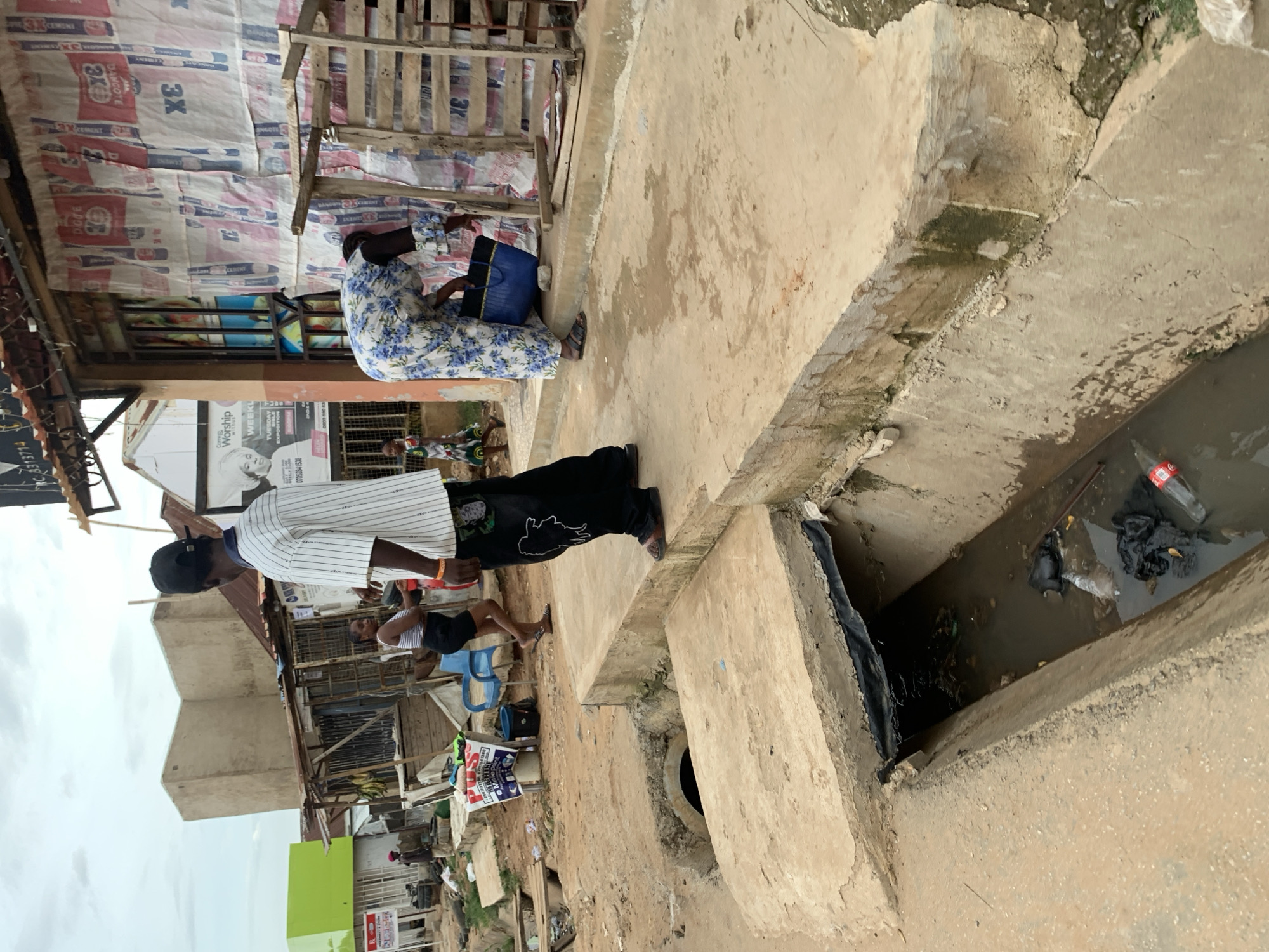 In Abuja, Nigeria, on a street with a concrete drainage channel, a man wearing a black baseball cap and a white and black striped t-shirt stands facing away from the camera. He is wearing black pants with a white design and brown sandals. Beside him, a woman in a blue and white floral dress is leaning down, holding a dark blue bag and interacting with a blue bucket. To the left, a young girl in a colorful dress walks away from the camera. In the background, a building with a corrugated metal roof is visible, along with advertisements and other structures. The drainage channel in the foreground contains murky water and debris, including plastic bags and a bottle.