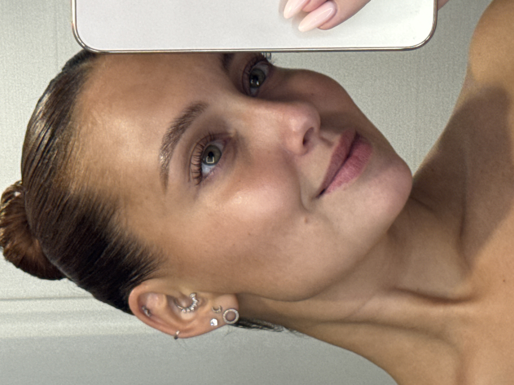 The image shows a close-up, profile view of a young woman's face, neck, and shoulder. She is looking towards the right of the frame, and part of a mobile phone is visible on the right side, held by her hand, which is also partially in the frame. The woman has her hair pulled back in a bun. She is wearing multiple earrings in her left ear. Her skin appears tanned, and she has a faint smile. The background is a plain, light-colored wall. There are no discernible landmarks or indicators to suggest a specific location within Chaikovsky, Russia.