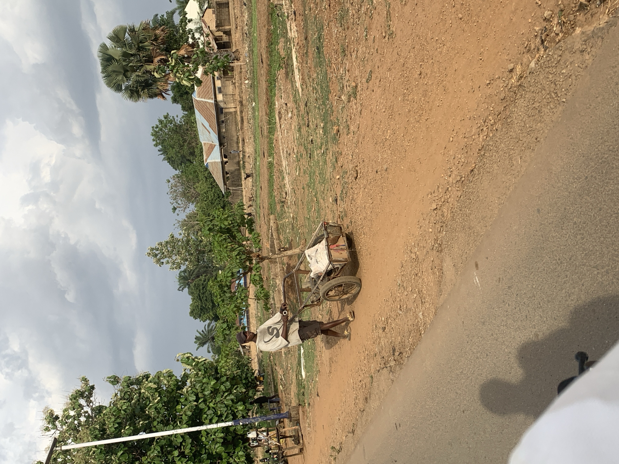 A man is pulling a two-wheeled cart along a dirt path in Maigizo, Nigeria. The man is wearing a light-colored shirt, dark shorts, and a baseball cap. The cart is partially filled with items covered by a white sheet. The path runs parallel to a paved road. The background includes trees, vegetation, and a building with a corrugated metal roof. The sky is overcast.