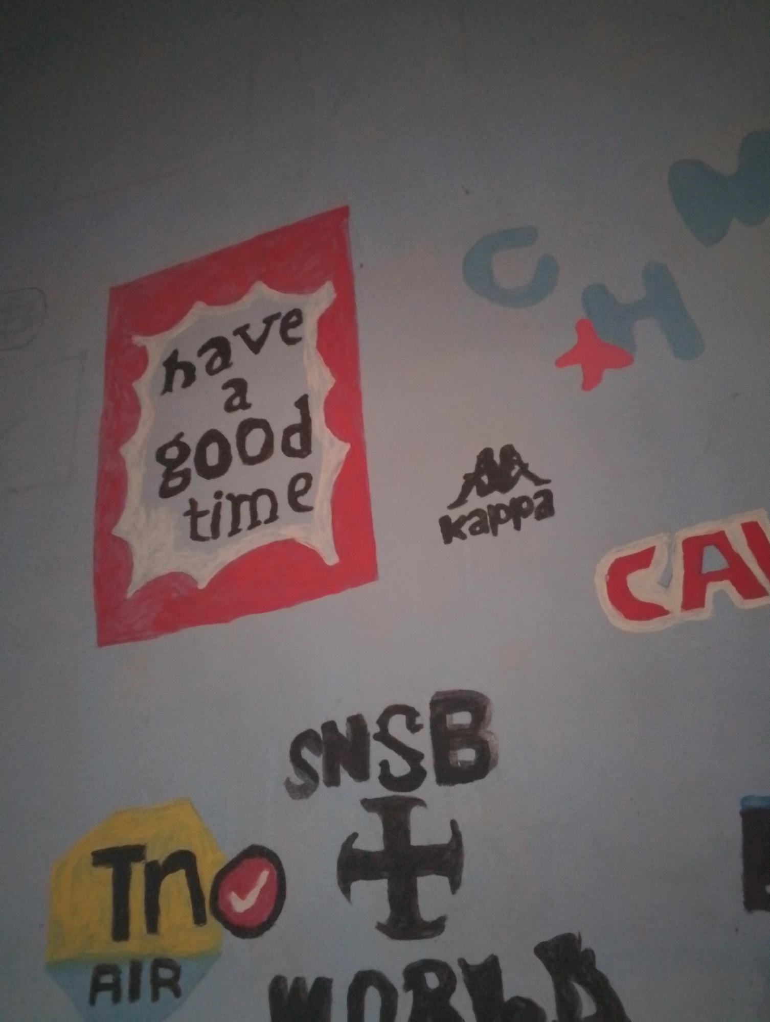 The image depicts a section of a wall with various hand-painted or drawn elements. Prominently featured is a red, scalloped-edged frame containing the words "have a good time" in dark, stylized lettering. To the right, there are scattered blue and red shapes, including what appears to be a star. Below these, a black Kappa logo with the text "kappa" is visible. Further to the right, a partially visible red and white logo reads "CA". In the lower left quadrant, a yellow and light blue shape displays "Tn" in black lettering above the word "AIR". Next to this is a red circle with a white checkmark. In the lower center, the letters "SNSB" are painted in dark, blocky letters, with a dark cross symbol directly below them. The bottom edge of the image shows more dark lettering, partially obscured, that appears to read "WORLD". The background is a light blue, textured surface, likely a wall. No specific city or location is identifiable from the image itself.