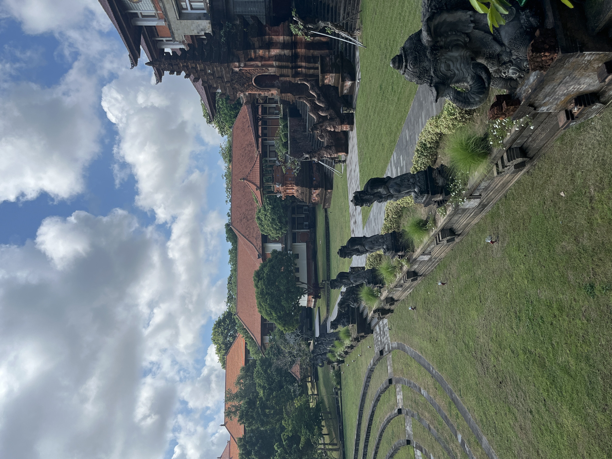 The image shows a wide view of an outdoor courtyard with a traditional Balinese architectural style, located in Kuta Selatan, Indonesia. The scene features a terraced grassy area on the left, resembling an amphitheater, with pathways winding through it. A long, ornate stone wall runs diagonally across the middle ground, adorned with statues of mythical creatures and carved balustrades. Beyond the wall, a paved pathway leads towards a large, intricately carved stone structure with a towering entrance and elaborate steps, typical of Balinese temple architecture. Several other buildings with traditional red-tiled roofs and white walls are visible in the background, surrounded by lush green trees. The sky above is partly cloudy with a mix of white cumulus clouds and blue sky. The overall atmosphere is serene and grand, showcasing cultural heritage and natural landscaping. There are no people visible in the image.