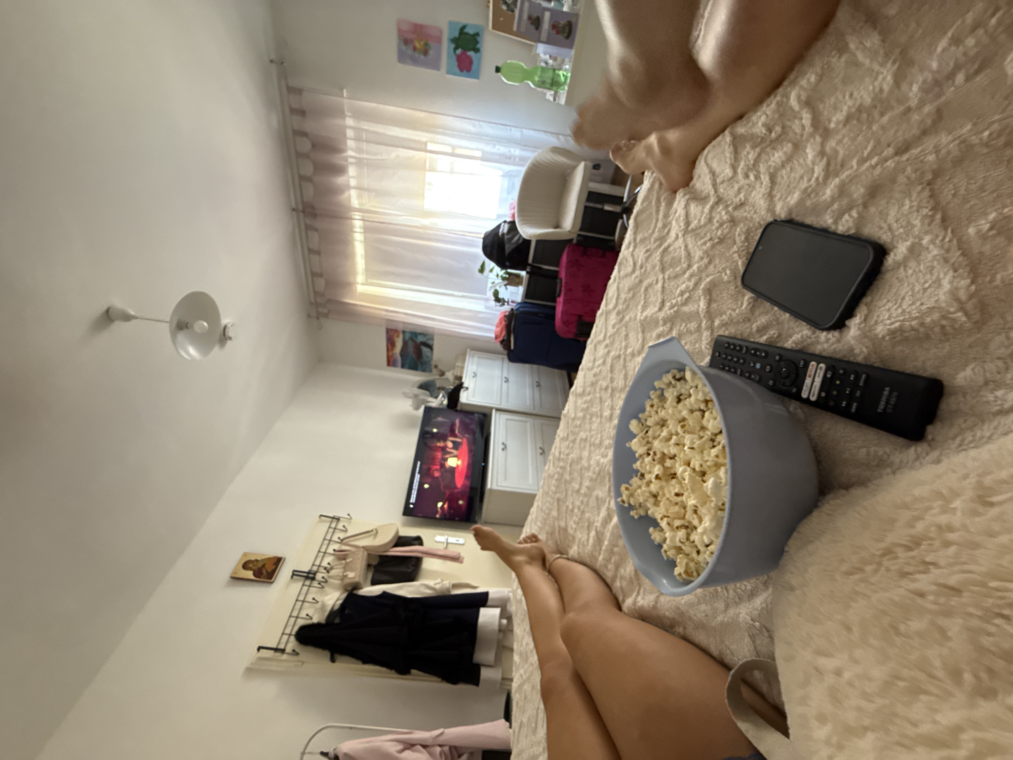 The image depicts a bedroom scene in Hanau, Germany. Two individuals, their legs visible in the foreground, are lying on a bed covered with a textured, beige blanket. A blue bowl filled with popcorn is positioned on the bed, next to a black television remote and a dark-colored smartphone. In the background, a white dresser with drawers is visible, with a television screen displaying a scene with two people. To the left of the dresser, a rack with hanging clothes and bags is present. Further back, sheer curtains partially obscure a window, allowing natural light to filter into the room. Artwork hangs on the wall, and a white chair and several pieces of luggage are situated near the window. A green plastic bottle and other decorative items are on a shelf. The ceiling features a white pendant light fixture.