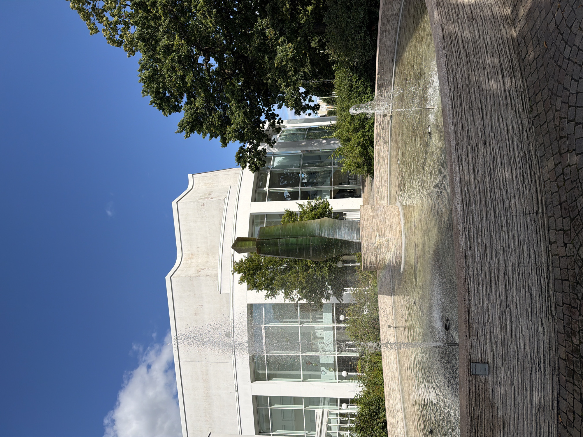 The image shows a fountain in front of a modern white building with large glass windows. The fountain has a stone base and a green glass sculpture in the center. Water sprays from multiple jets in the fountain. Lush green trees and bushes surround the building and fountain. The sky is blue with scattered white clouds. The scene appears to be outdoors on a sunny day. This is the fountain located at the entrance of Växjö Konserthus (Växjö Concert Hall) in the city of Växjö, Sweden. There are no people visible in the image.