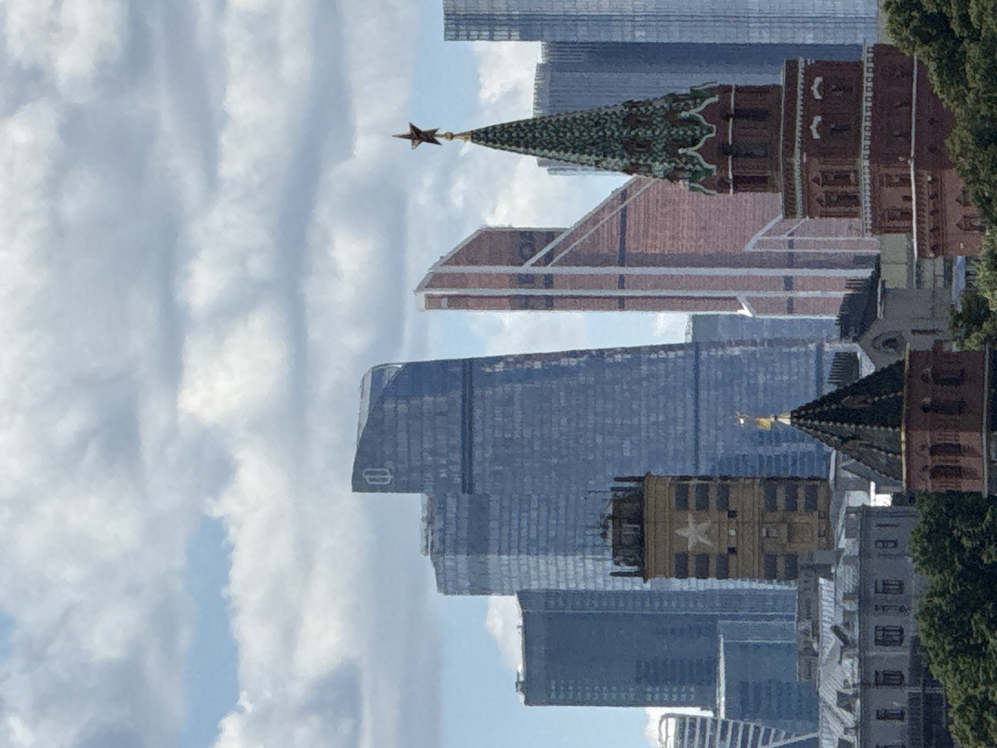 The image shows a cityscape with modern skyscrapers and traditional architecture in Moscow, Russia. In the foreground, there are two towers, one made of brick with a green, tiered roof topped with a star, and another more square-shaped tower with horizontal dark stripes and a white star emblem. Behind these towers, several modern skyscrapers are visible, including tall, glass-fronted buildings with distinctive architectural shapes. One skyscraper on the left is tall and blue, while another to its right has a unique angled design in reddish-brown. The sky is partly cloudy with blue sky peeking through. Green trees are visible at the bottom of the image, suggesting a park or green space within the urban environment. The scene captures a blend of historic and contemporary architectural styles characteristic of Moscow.