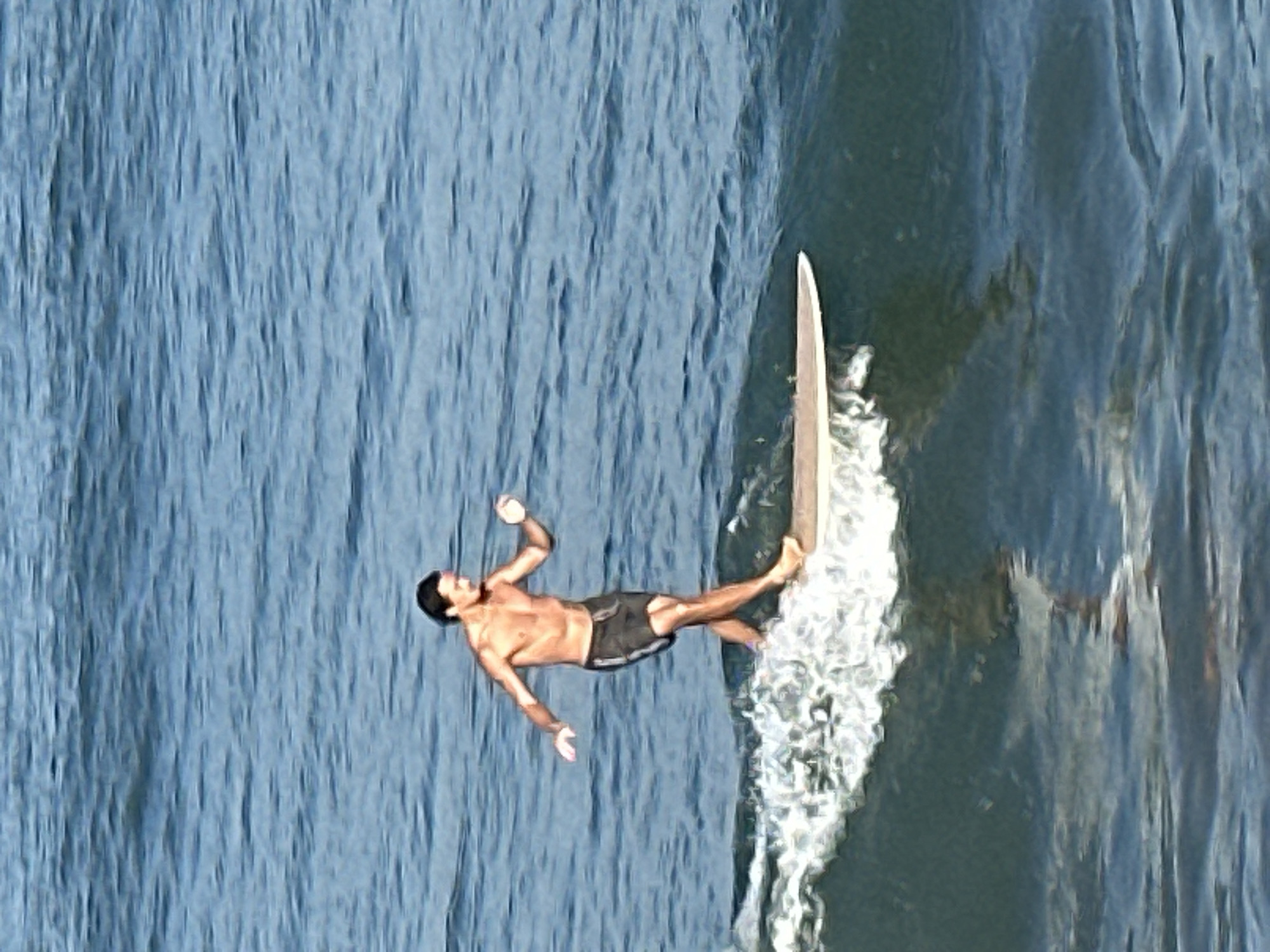 In the azure waters off the coast of North Kuta, Indonesia, a lone surfer rides a wave. The surfer, a man with dark hair and a tan physique, is shirtless and wearing dark board shorts. He stands on a light-colored surfboard, his arms outstretched for balance as he glides across the water. The wave he is riding is breaking, creating a foamy white crest that surrounds the board. The ocean is calm with gentle ripples, reflecting the clear blue sky above. The overall scene captures a moment of serene athleticism amidst the natural beauty of the Indonesian coastline.