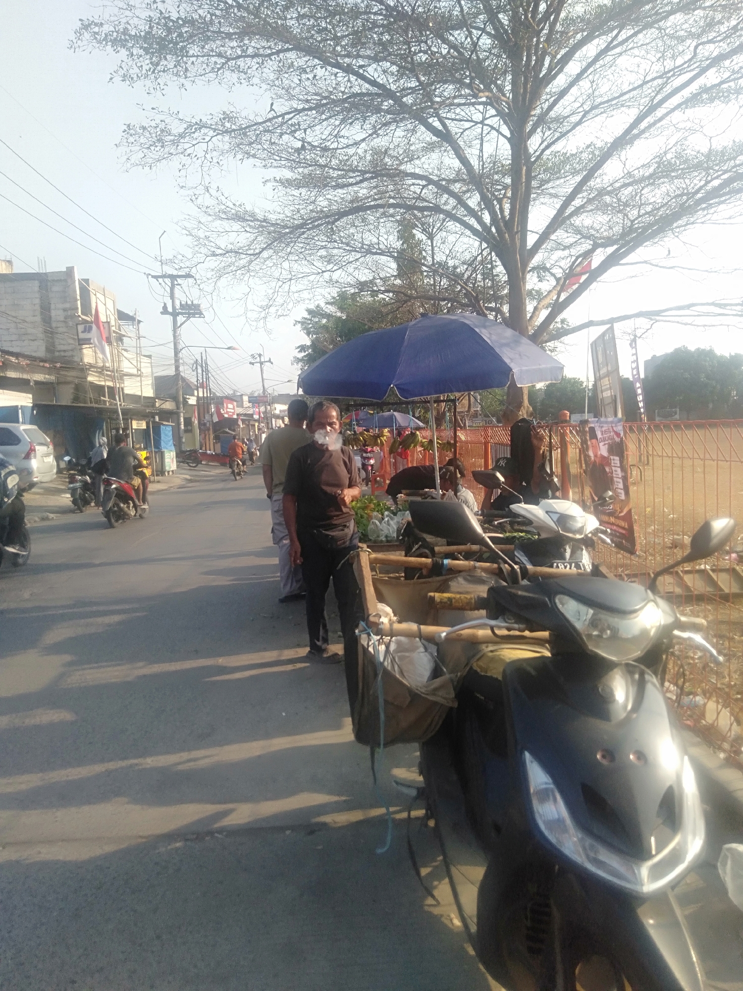 A street scene in Sukadami, Indonesia, captures a bright, sunny day with long shadows cast across the asphalt. In the foreground, a dark-colored scooter with a silver headlight and handlebars is parked. It has a brown woven bag attached to its front. Further down the street, a man wearing a dark t-shirt and black pants stands with smoke emanating from his mouth, suggesting he is smoking. He is facing towards the right of the frame. Next to him, another person in a light-colored t-shirt and grey pants stands in profile. Behind them, a blue umbrella shades a stall displaying produce, with more people visible in the background. A tall tree with bare branches dominates the upper portion of the image, with power lines crisscrossing the sky. Buildings line the left side of the street, and several other motorcycles are parked or in motion. A red and white Indonesian flag is visible on one of the buildings.