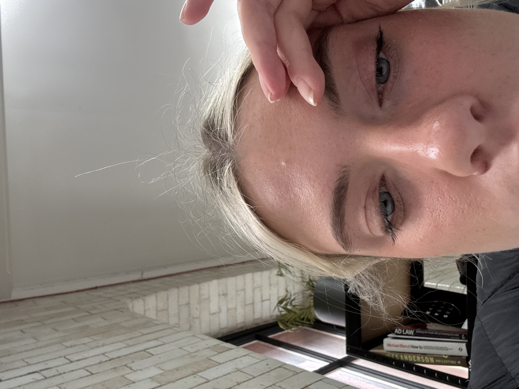 A close-up, low-angle photograph captures the upper portion of an adult female's face, her gaze directed downwards. She has blond hair with visible darker regrowth at the scalp, and blue eyes, with black eyeliner applied to the upper lash line of her visible right eye. A small blemish is present on her forehead. Her left hand, showing visible fingernails, rests with two fingers lightly touching her forehead above her left eye. She is wearing dark, possibly quilted, apparel.
The background reveals an interior setting. To the upper left, a light-coloured brick wall, possibly whitewashed, runs diagonally upwards, featuring a vertical reddish-orange pipe. This wall adjoins a window with dark framing. Through the window, a potted plant with green foliage is visible on a dark shelf or ledge. Beneath the window, a black metal shelving unit contains several books, with discernible titles including "AD LAW" and a partially visible "Michael Bond: How to...". The upper right portion of the image shows a plain white ceiling. The exposed brickwork and industrial-style window frames are consistent with architectural features found in renovated urban structures, commonly seen in areas such as Ancoats or the Northern Quarter within Manchester, United Kingdom. No specific address or definitive landmark is identifiable.