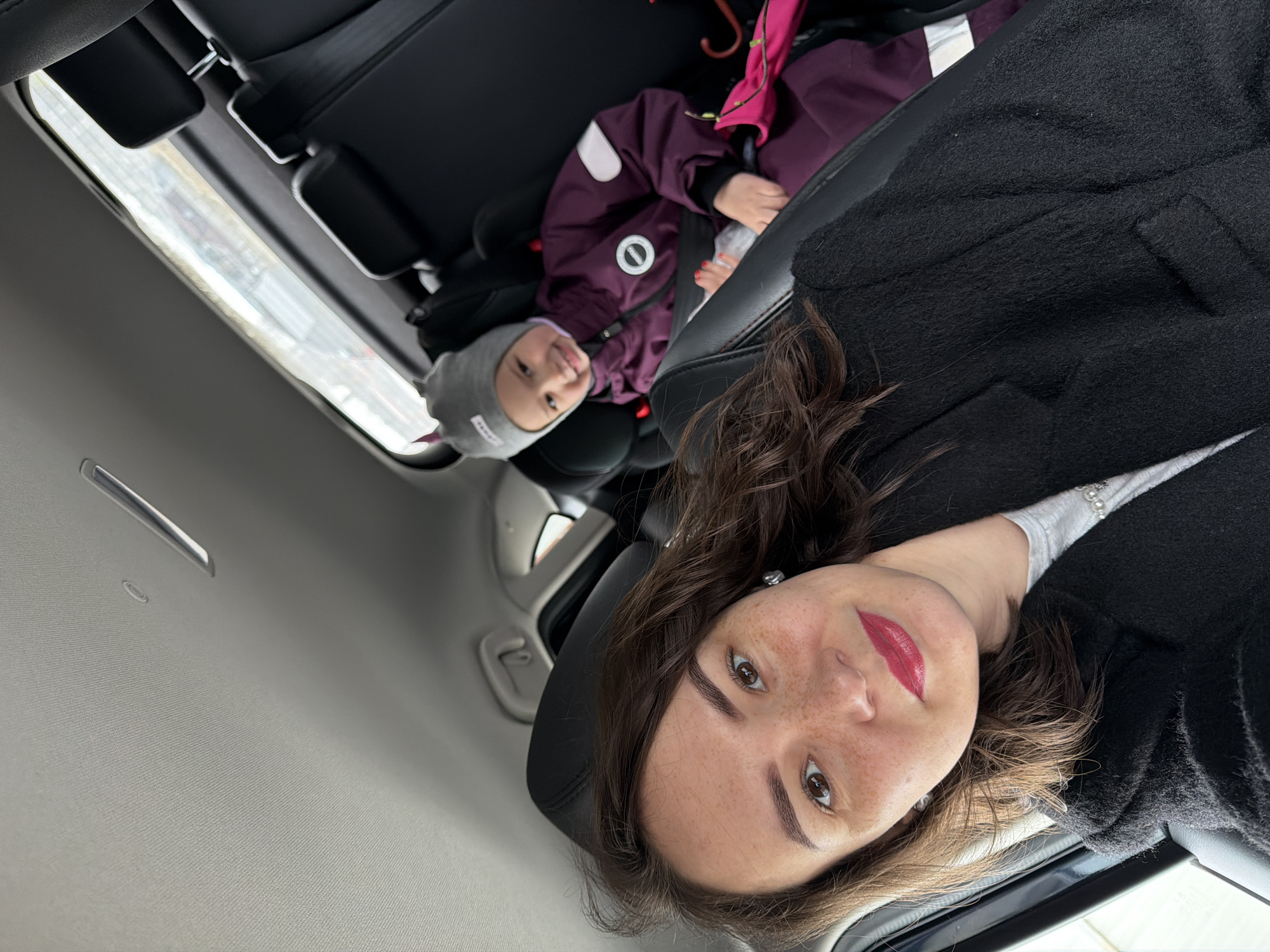 A woman and child are seated in the interior of a car. The woman is facing the camera and partially obscured by her dark-colored jacket. She has light skin, brown hair, and is wearing lipstick. A child, wearing a gray knit hat and a purple jacket, is visible in the back seat. The car's interior is visible, including the headliner and a sunroof. The location is Chaikovsky, Russia, inferred from the exterior view through the sunroof. The angle of the shot suggests the woman may be taking a selfie.