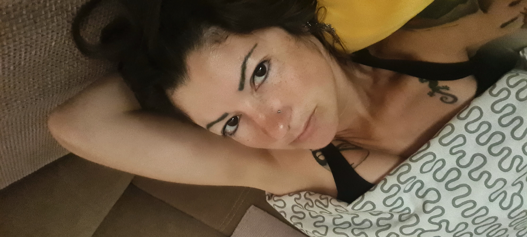A person with dark hair and fair skin lies on a surface, partially covered by a white and grey patterned blanket. The person has a nose piercing and visible tattoos on their chest and shoulder. The background consists of a yellow pillow and a textured surface. The location is not discernible from the image.