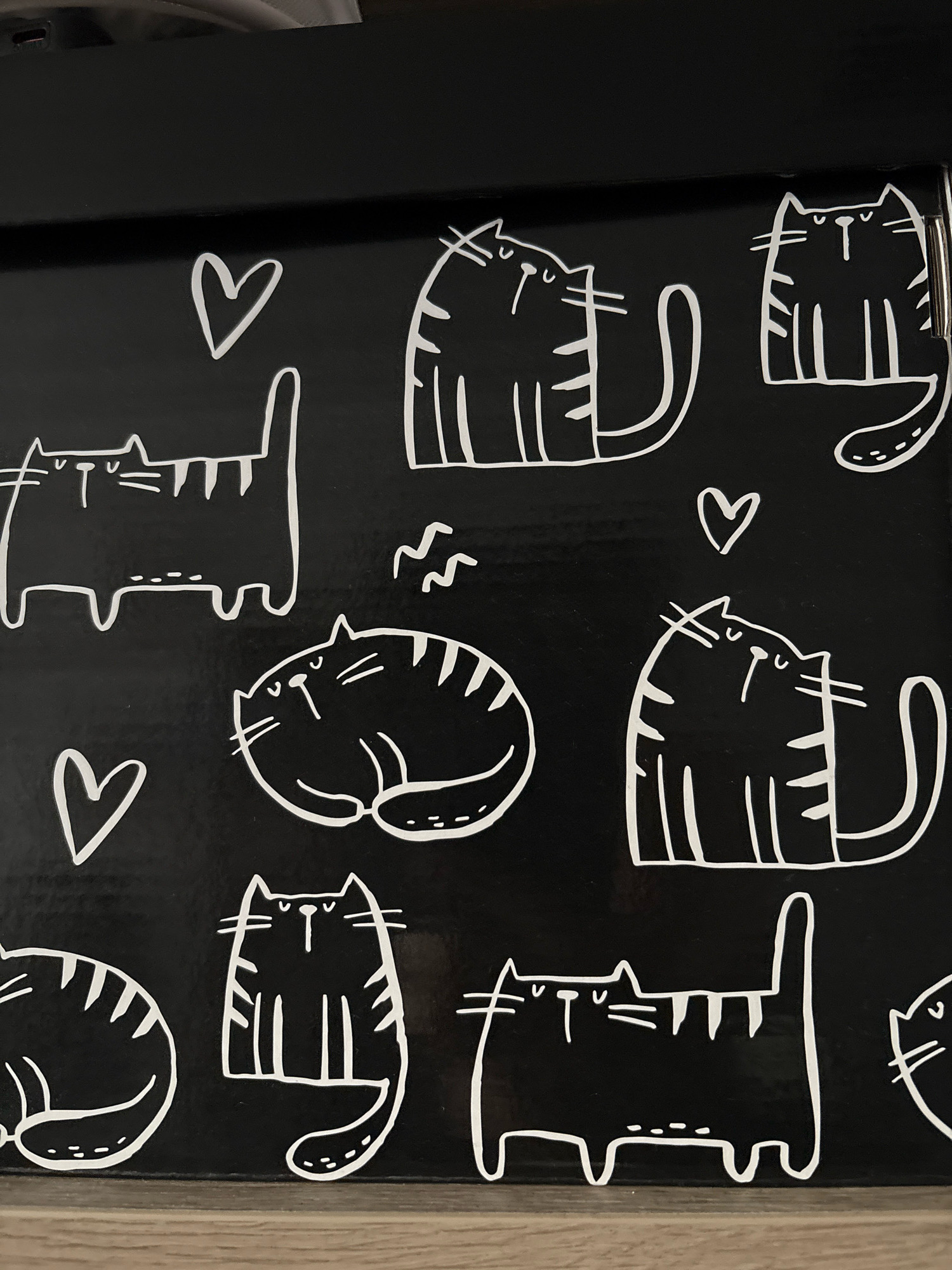 The image shows a black surface with a repeating pattern of white line drawings of cats and hearts. There are at least six distinct cat illustrations, varying in pose and detail, with some featuring stripes. A few white hearts are also scattered among the cat designs. The surface appears to be a flat, dark material, possibly part of a box or furniture. The lighting suggests an indoor environment. There is no visible information in the image to determine a specific location in Zapadny, Russia, or any other city.
