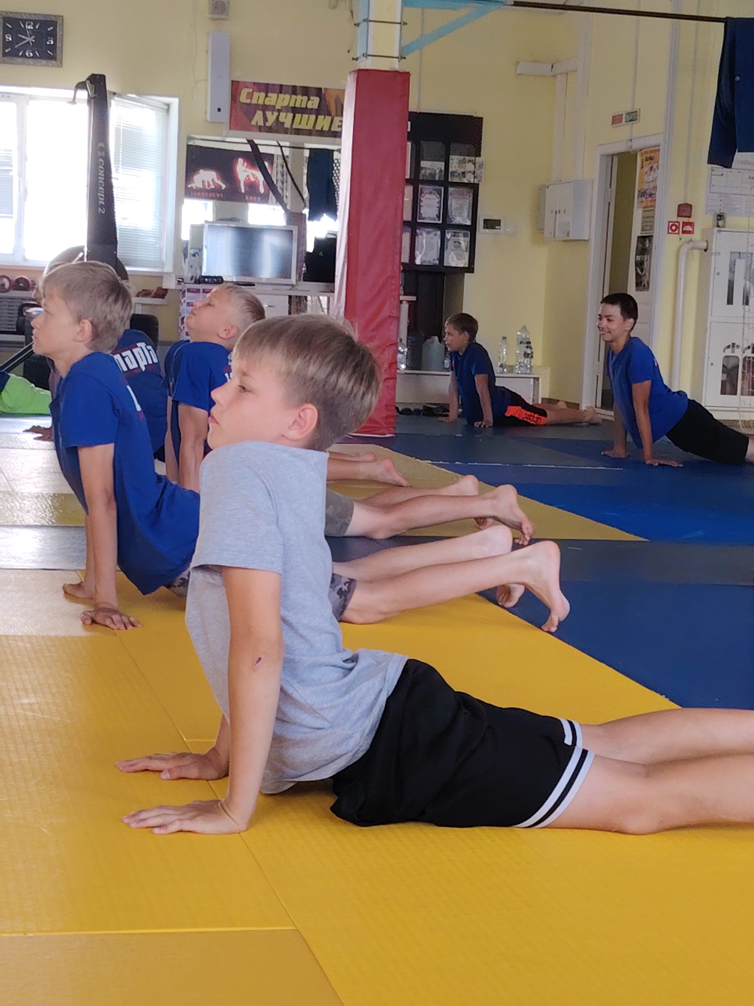 The image shows a group of young boys in a gym, possibly a sports hall, participating in a physical exercise class. The floor is covered with yellow and blue mats. Several boys are in a cobra pose, with their upper bodies arched off the mat. In the background, there are posters on the wall, including one with Russian text that translates to "Sport for All." A rowing machine is visible near a window. The overall environment suggests a sports training facility. The specific location in Chaikovsky, Russia, is indicated by the text on one of the posters.