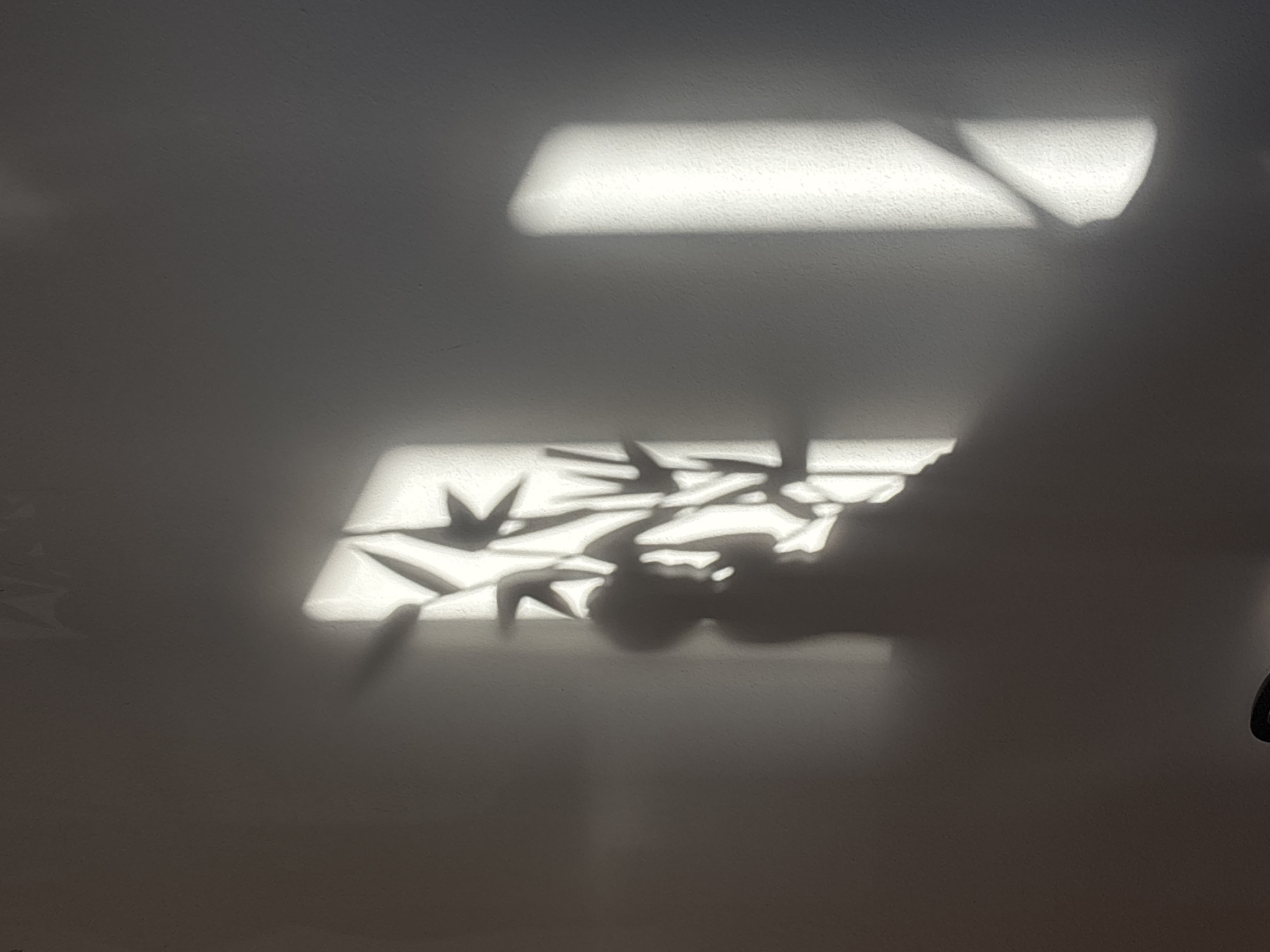 The image shows a shadow of a person sitting on the floor next to a window with blinds. The blinds cast a patterned shadow on the wall. The image is taken in an indoor setting, likely a room in Budva, Montenegro, judging by the context provided. The lighting suggests it is daytime.