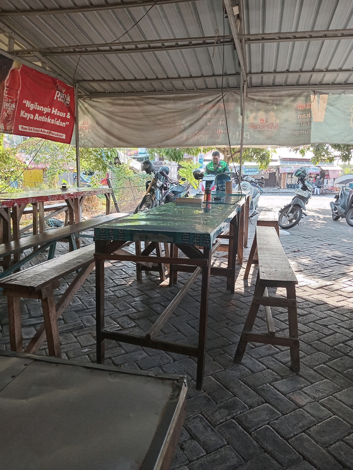 The image depicts an outdoor seating area of a food stall or small restaurant in Kembangan, Indonesia. The foreground shows a rustic wooden table with a green tiled surface, accompanied by wooden benches. Behind this table, another longer wooden table with a red top is visible, also with benches. The ground is paved with interlocking brick pavers. In the background, several motorcycles are parked, suggesting a common mode of transportation for customers. A man in a green shirt is standing near the tables, possibly attending to customers. Various banners are hung overhead, one of which, a red banner, displays text that appears to be an advertisement for "Pucuk Harum," a drink. Another banner behind it is partially visible and appears to have text in a light color. The overall setting suggests a casual, open-air dining establishment situated along a street or pathway, likely in a moderately busy area given the presence of parked motorcycles and the presence of shops in the distance. Sunlight casts shadows on the ground, indicating it is daytime.