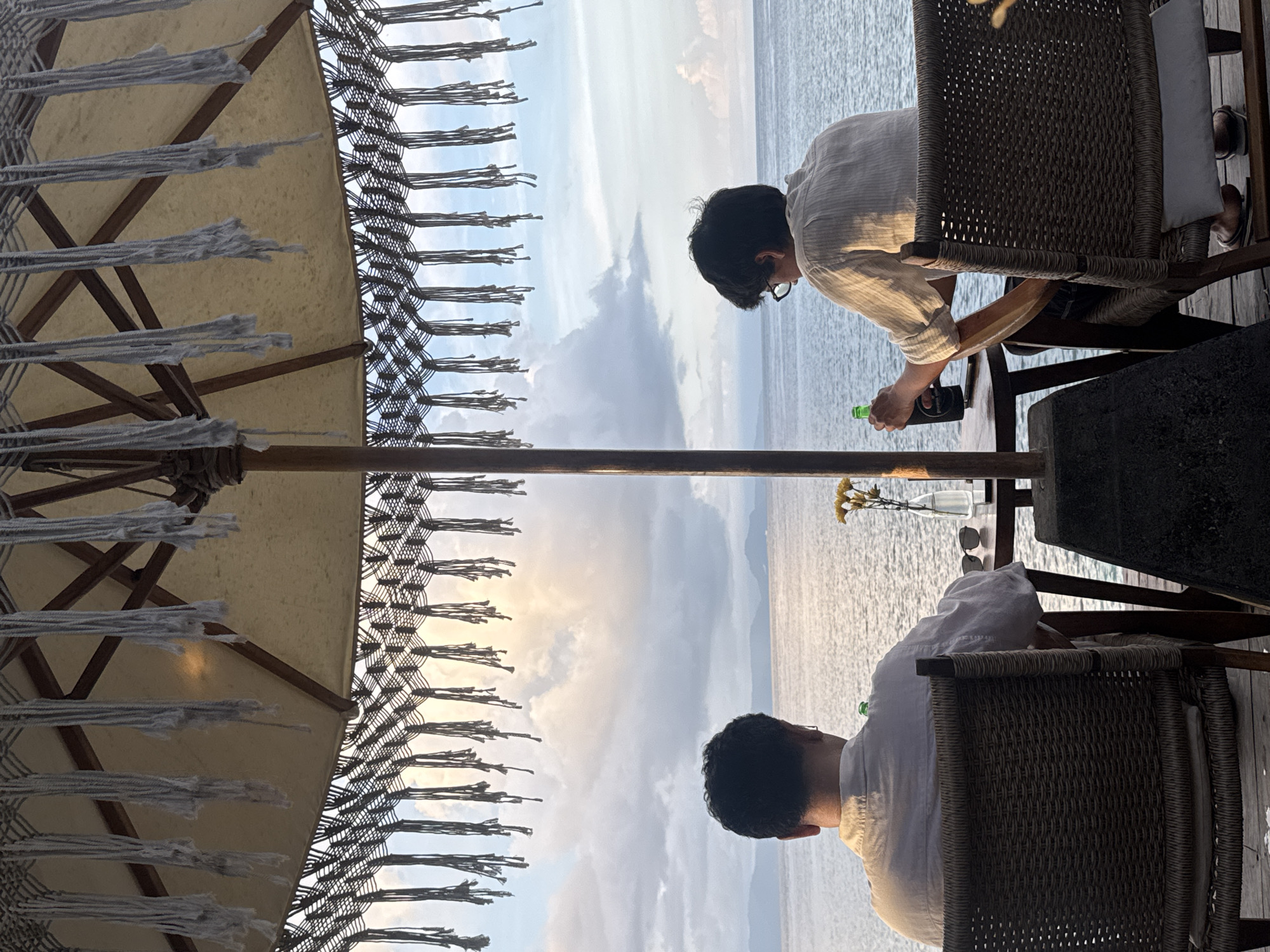 The image captures a person, presumably an adult, dressed in light-colored clothing, possibly a shirt or blouse. They are seen seated in a wicker chair at an outdoor establishment. In their hands, they appear to be handling or pouring from a green bottle. On the table next to them, there are flowers in a vase and a pair of sunglasses. The foreground is dominated by a fringed sunshade, likely part of an umbrella or awning. Beyond the sunshade, the background reveals a seascape with a view of the ocean and distant landmasses, suggesting an elevated vantage point. Given the reference to Nusa Penida, Indonesia, it is likely that the photo was taken at a coastal venue there, possibly a restaurant or lounge with ocean views. The lighting indicates either early morning or late afternoon.