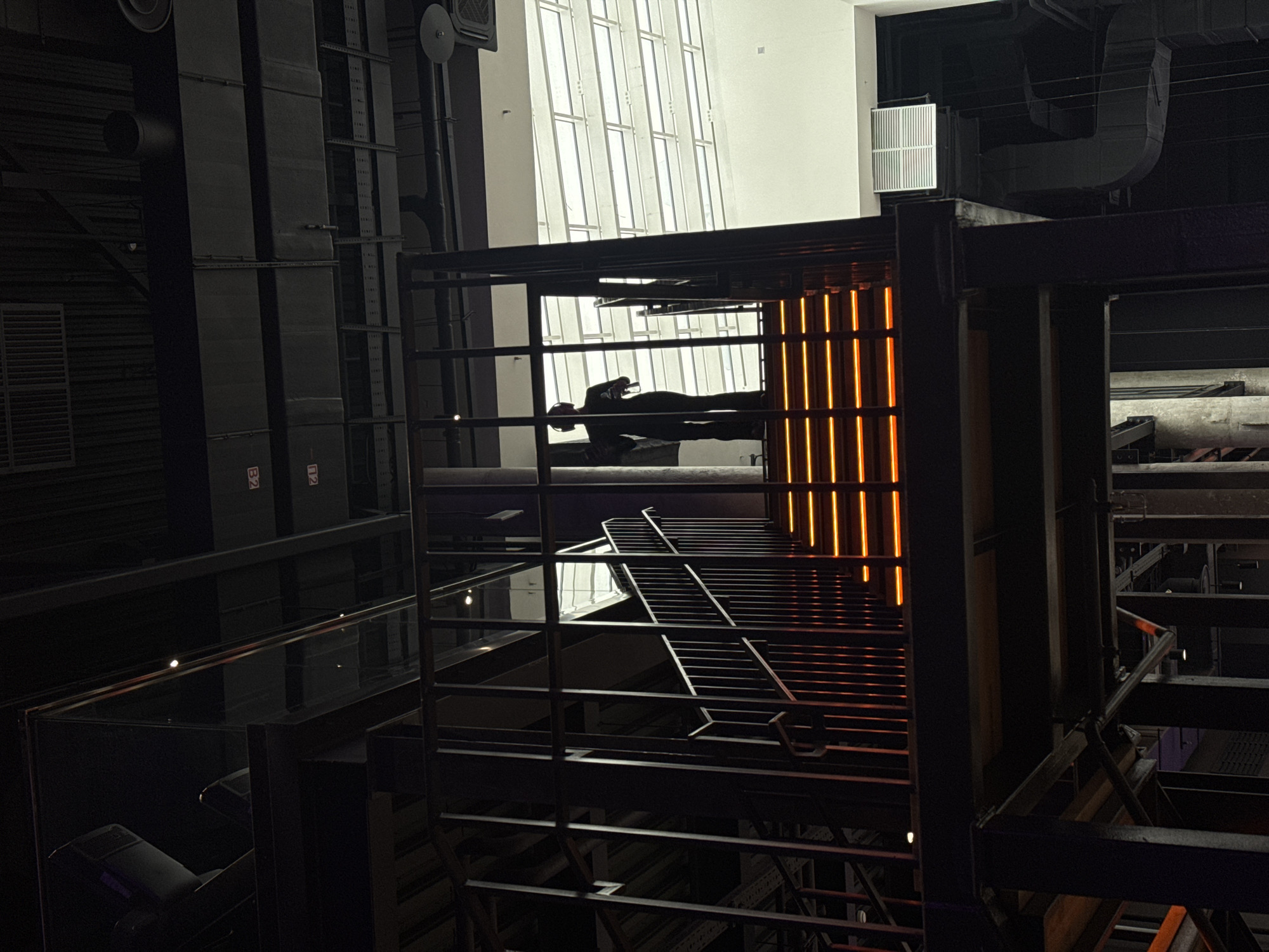 This is a dark, vertically oriented photograph capturing a modern architectural interior, possibly an industrial-chic space or a multi-level hall. The scene is dominated by a complex network of dark metal frameworks, including girders, beams, and railings, that create distinct levels and enclosures.
In the upper-middle section of the image, a human figure is prominently featured, lying horizontally within one of these framed structures. The person is silhouetted against a large, brightly lit windowed wall in the background, which displays a grid pattern of white frames. They appear to be holding a small object, possibly a phone or a drink, near their face.
To the immediate right of the reclining figure, a striking vertical element comprises numerous glowing orange light strips, providing a warm, vibrant contrast against the otherwise dark palette of the scene. This illuminated structure extends downwards into what appears to be a tiered seating area or a staircase with horizontal slats.
Various dark, industrial elements such as large ventilation ducts and pipes are visible in the upper-left and upper-right portions of the image, blending into the shadowy background. A white rectangular vent with vertical grates is also discernible in the upper-right. Reflective surfaces, possibly glass panels, are present on lower levels, catching subtle glints of light.
On a dark vertical column in the upper-left, red text "B2" and "N2" is visible. The overall atmosphere is modern and somewhat dramatic, with strong contrasts between light and shadow, suggesting an evening or nighttime setting indoors. The location is Moscow, Russia.