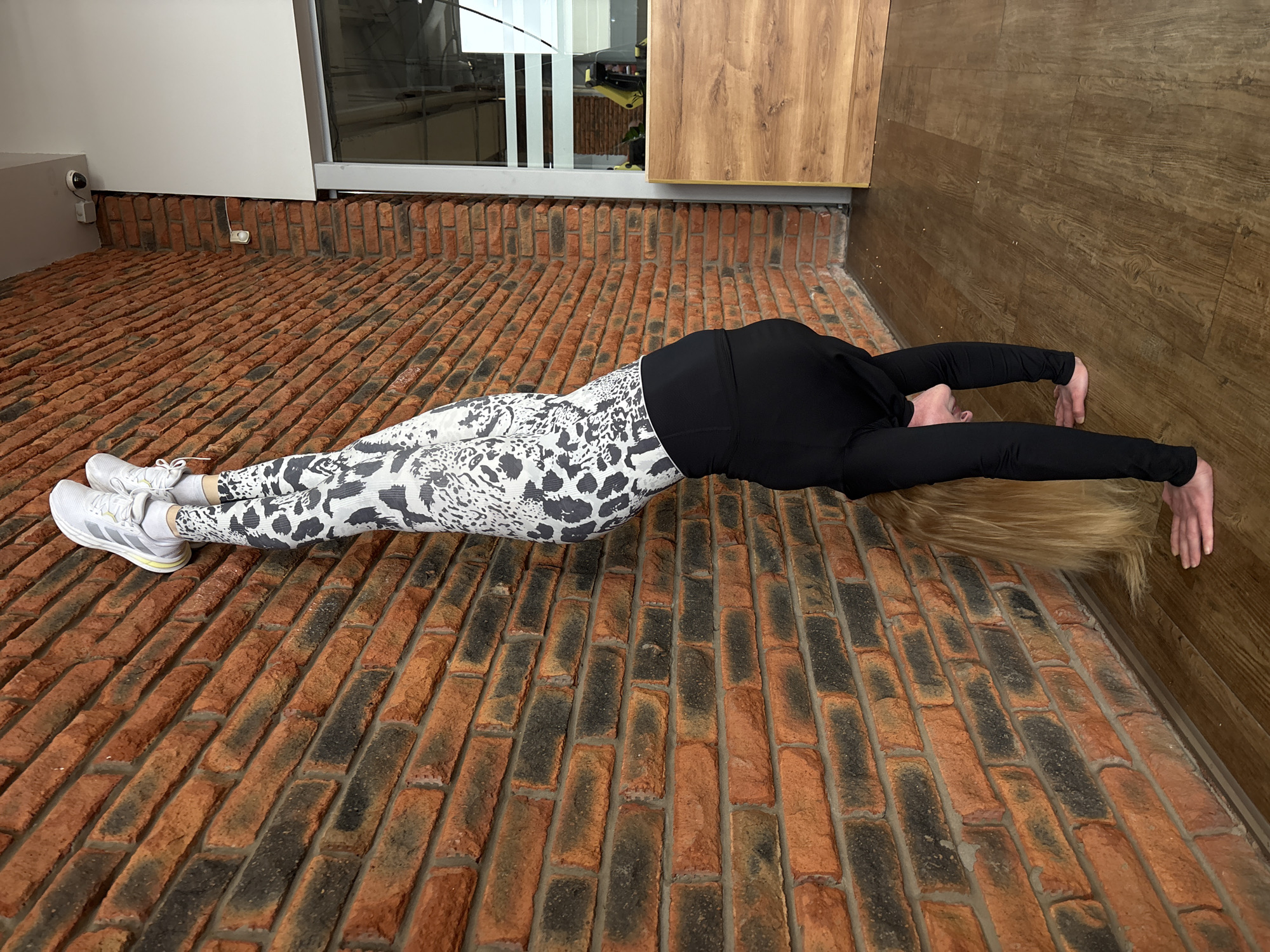 An indoor scene captures a person engaged in a stretching exercise on a textured brick floor. The individual, positioned in a backbend on their stomach, extends their arms forward, pressing their hands against a vertical wooden-paneled wall. Their long blonde hair cascades onto the floor, obscuring their face. They are dressed in a black long-sleeved top and white leggings adorned with a black abstract or animal print pattern, complemented by white athletic shoes with gray accents.
The floor is made of reddish-brown bricks, many featuring darker, almost burnt-orange patches, laid in an angled pattern. The wall to the person's right is covered in wooden panels. To the left, a plain white wall is visible, featuring a small black security camera and a white electrical socket mounted near the brick floor line. Above this, a large window or glass partition reflects an interior space, likely another room or objects within the building. Adjacent to the window on the right is a wooden cabinet or wall section.
The bright lighting suggests it is daytime. The setting combines industrial-style brickwork with modern white walls and warm wooden features, indicating a contemporary indoor environment, possibly a fitness area or a uniquely designed interior space in Chaikovsky, Russia. No discernible text is visible within the image. The person's posture suggests concentration and physical activity.