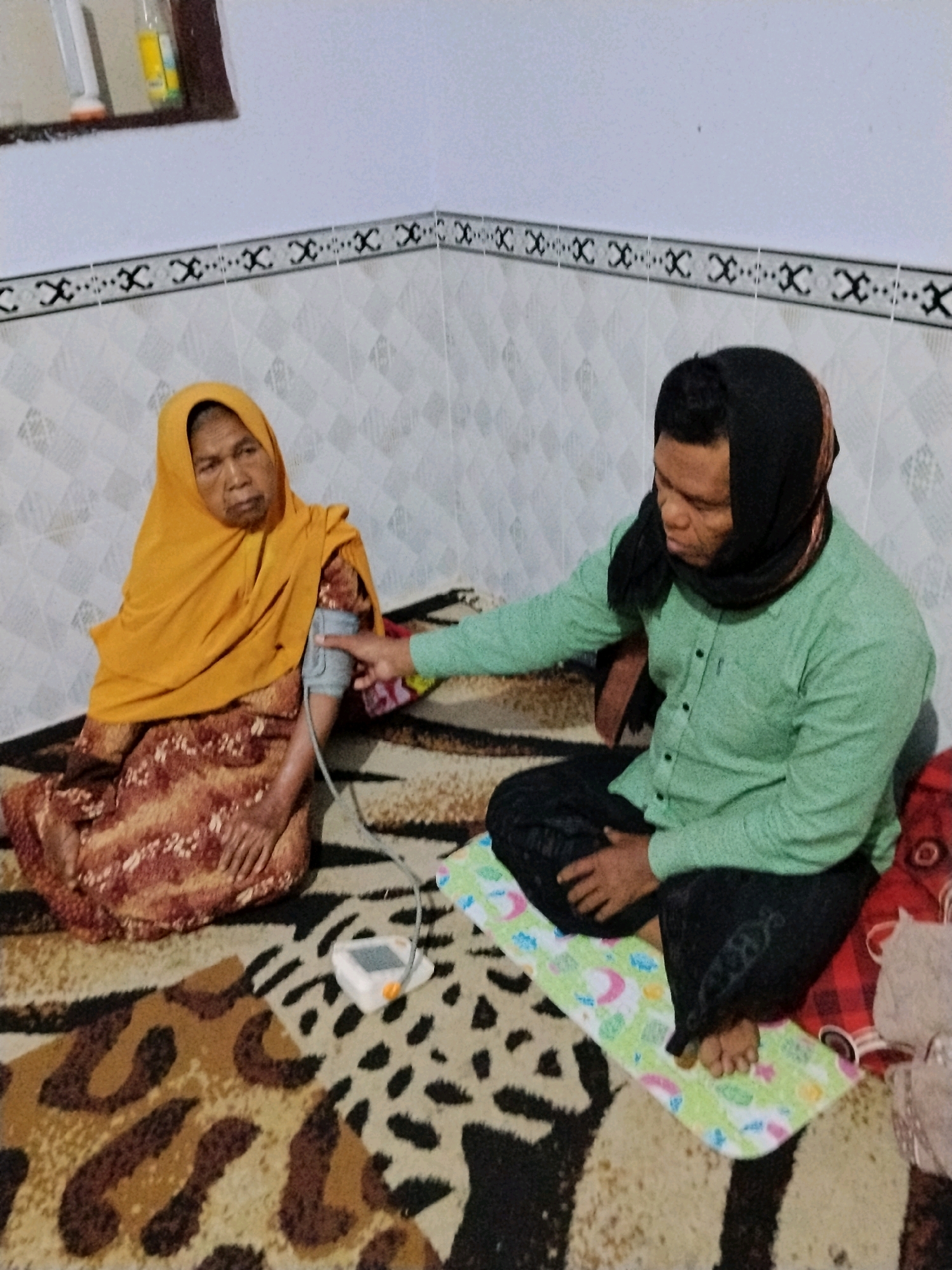In the city of Panggungan, Indonesia, an older woman wearing a yellow headscarf and a patterned dress sits on a leopard-print rug. A man, also on the rug and wearing a green shirt and a black headscarf, is taking her blood pressure. A white blood pressure monitor with an orange button is visible on the rug between them. The scene appears to be indoors, with tiled walls and a patterned border along the top.