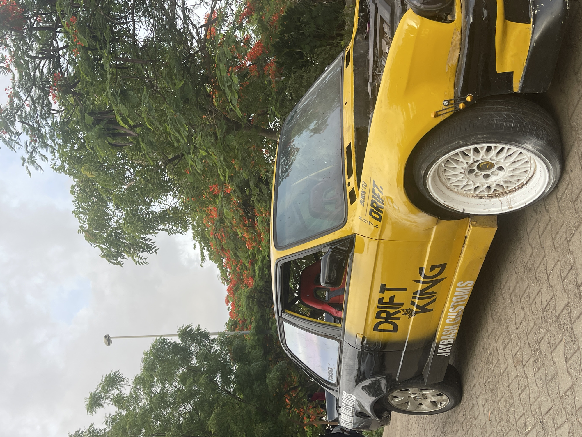 A heavily modified yellow and black car with "DRIFT KING" and "JAYBASH CUSTOMS" decals sits on a paved surface. The car is parked outdoors near trees with red flowers. A streetlight is visible in the background. The location is Abuja, Nigeria.