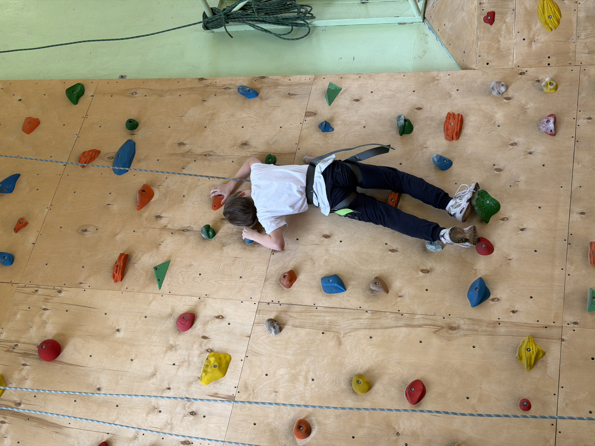 A child wearing a climbing harness, white shirt, and dark pants is lying on a climbing wall. The wall is made of light-colored wood and has numerous colorful climbing holds attached to it. A rope is strung horizontally across the bottom of the wall. In the upper left corner, a coil of rope is visible against a light green wall. The location is possibly in Izhevsk, Russia, as implied.