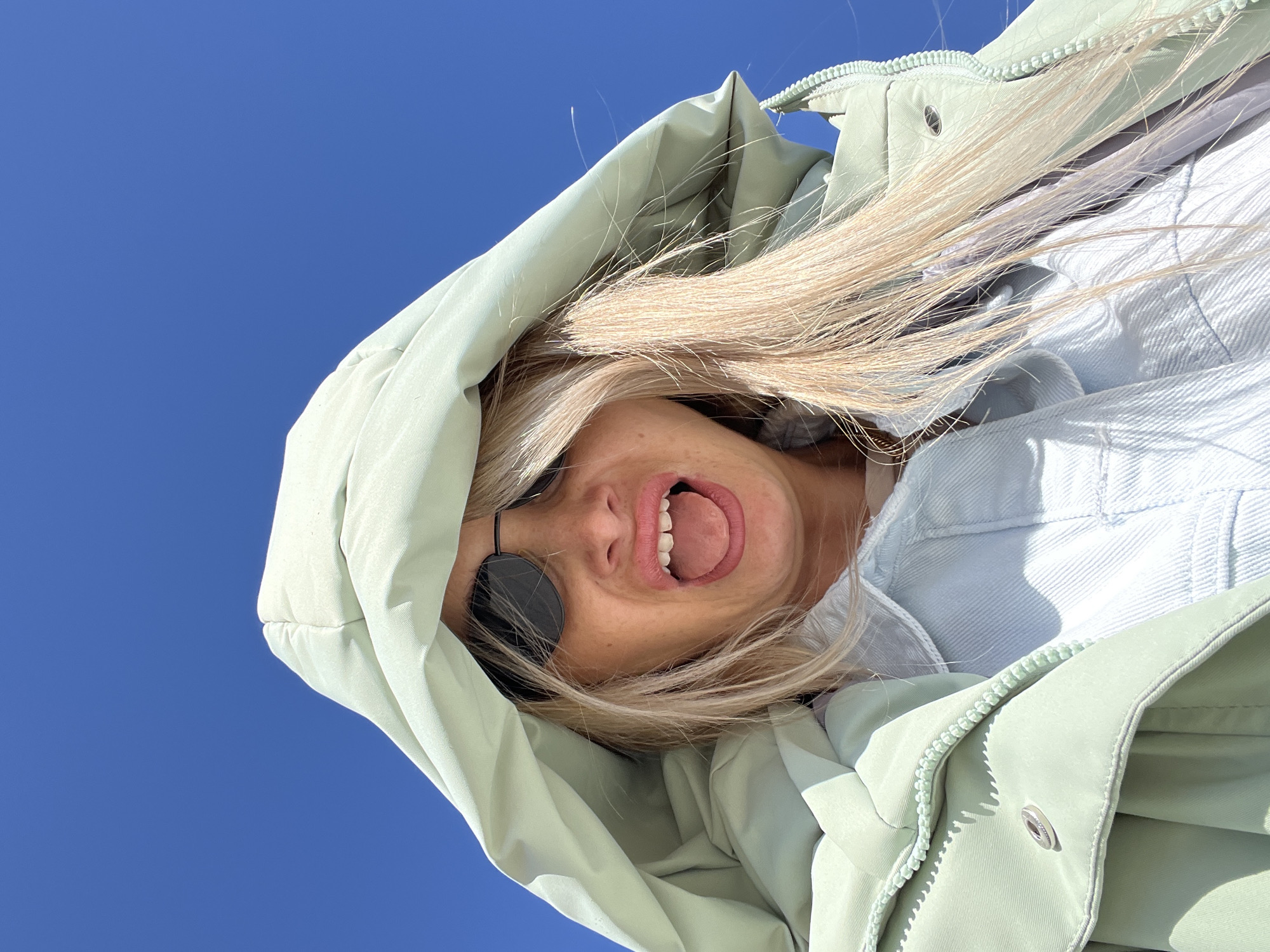 A low-angle shot of a blonde woman in a light green hooded puffer jacket, set against a clear blue sky. She wears round sunglasses and has her mouth open. A light blue denim jacket is visible beneath the puffer jacket. This image does not contain enough information to determine the location or city it was taken in, nor is it able to specify any actions or interactions.