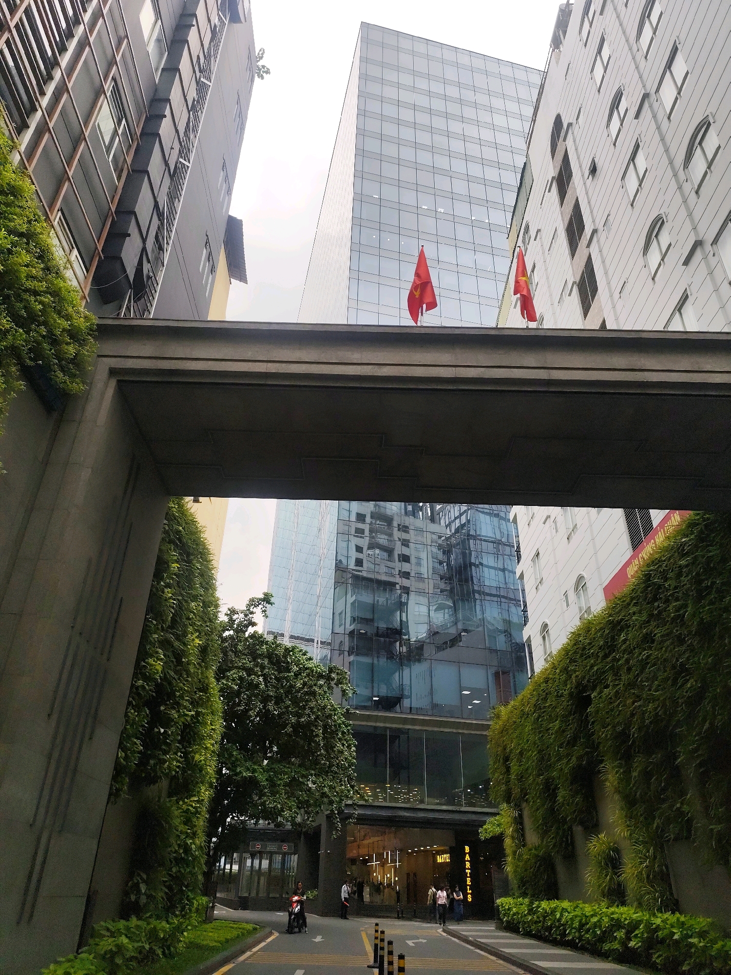 This is a street-level, wide-angle photograph taken in Ho Chi Minh City, Vietnam, looking up at the architecture of the city. The foreground features a large concrete overpass draped with lush green ivy, creating a tunnel-like effect. Below the overpass, a street curves to the right, with a motorcycle rider visible in the middle of the road. Two individuals stand on the right sidewalk.
Beyond the overpass, several modern buildings dominate the frame. To the left, a dark building with a grid-like facade rises. In the center, a tall, contemporary glass skyscraper reflects the sky and surrounding structures. To the right, a lighter-colored building with arched windows stands. Two Vietnamese flags, red with a yellow star, are prominently displayed on the roofline of the central glass building. The sky is overcast. The scene conveys an urban environment characterized by a mix of older and newer architectural styles, with abundant greenery integrated into the built landscape.