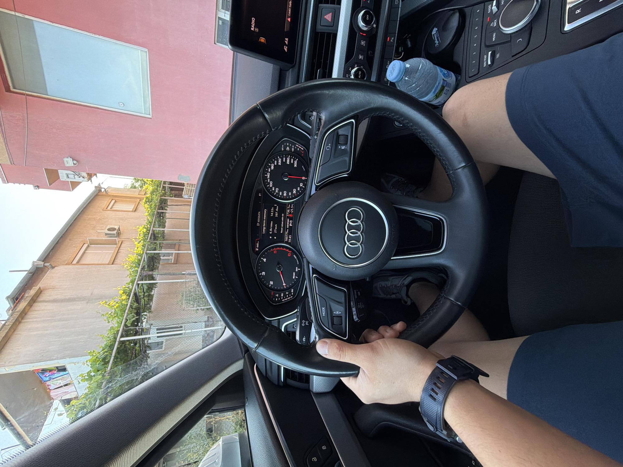 An interior shot captures a person's hands on the steering wheel of an Audi car, parked in Burgas, Bulgaria. The driver, wearing dark blue shorts and a wristwatch, is seated behind the wheel, with their feet visible in grey sneakers. The car's dashboard displays various indicators and a touch screen. Outside the car, a pink building with a large window and an adjacent beige building with balconies are visible. Green vines are growing along a fence in front of the buildings, and laundry hangs on a balcony. The scene suggests a sunny day in an urban environment.