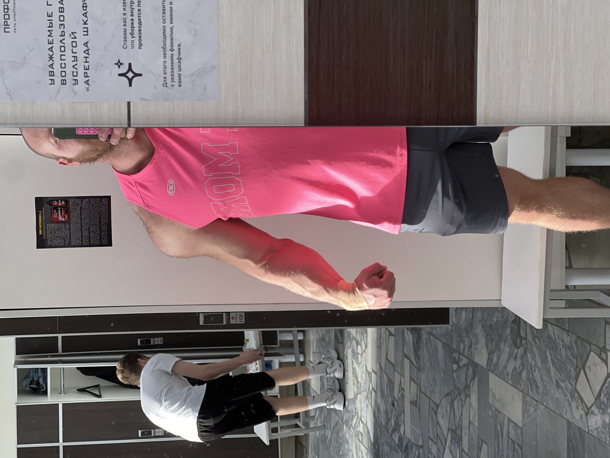 The image shows a man taking a selfie in front of a mirror in what appears to be a locker room or changing area. The man is wearing a pink tank top and gray shorts, and is flexing his arm. Behind him are lockers and another man standing near them. In the mirror, a sign in Russian is visible, which states, "Уважаемые гости, воспользуйтесь услугой 'Аренда шкафа'," which translates to "Dear guests, use the 'Locker Rental' service." The sign also provides instructions on leaving information for locker cleaning. The location is likely in Chaikovsky, Russia.