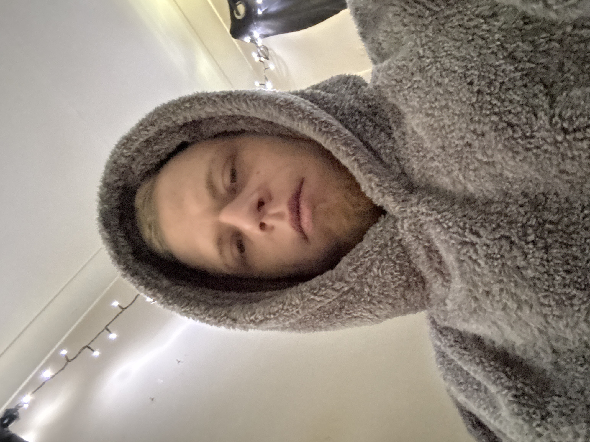 A selfie shows a person with fair skin and blonde hair, partially covered by a large, gray, fuzzy hooded garment. The person's face is angled slightly downward, with the nose centered in the frame. Below the person, a white wall is visible with a string of small, white lights along the wall's edge. The location is inside a building in Great Yarmouth, United Kingdom, suggested by the person's dress and decor. The subject is likely taking a selfie indoors in a residential space. The person appears to be looking at the camera.