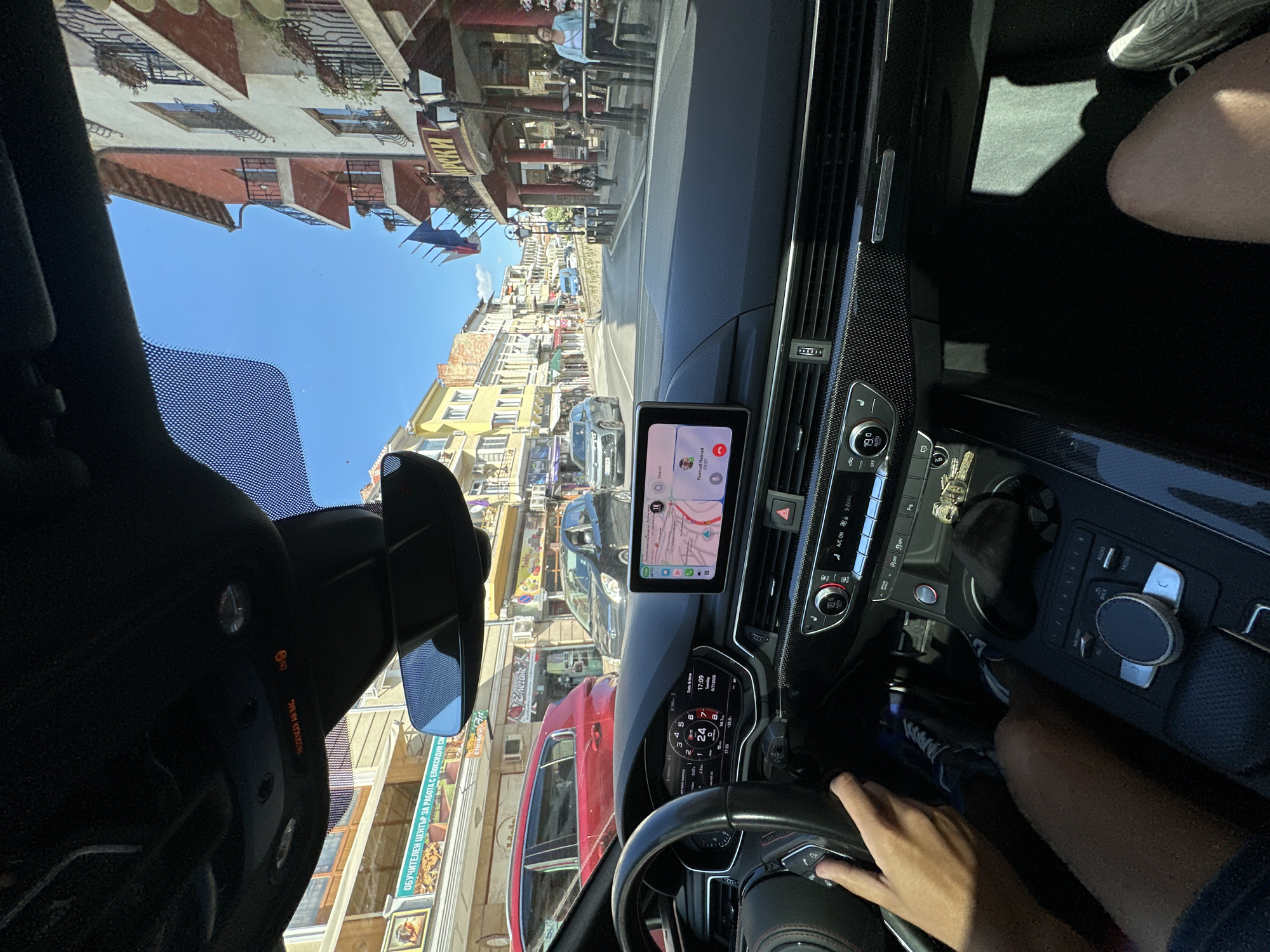 The image captures a first-person perspective from the driver's seat of an Audi (likely an S5/RS5 based on the interior details and carbon fiber trim), looking out onto a sunny street in Veliko Tarnovo, Bulgaria.
Inside the car, the driver's hands are positioned on a black leather steering wheel with red stitching. The virtual cockpit display shows a speed of '0', a date of '6/7/2026', and a time of '17:09'. The central infotainment screen displays a map application and an active phone call from "Преслав Пейчев" (Preslav Peychev). Carbon fiber trim extends across the dashboard, marked with a "Quattro" badge on the passenger side. The climate control system indicates a temperature setting of '19.0' degrees Celsius and 'AUTO' mode. The center console features an MMI controller and a gear selector, with a golden-colored charm attached to a key fob. Above the rearview mirror, an overhead panel displays "PASSENGER AIR BAG" with an illuminated 'ON' indicator. The driver's left leg, clad in shorts, is visible in the lower left, while the passenger's right leg and a grey/white striped shoe are visible in the lower right.
Outside the car, the scene depicts a narrow, asphalt road that curves slightly to the right and appears to ascend a gentle incline under a clear blue sky. On the left side of the street, buildings with lighter facades house shops. A prominent sign in Bulgarian reads "ОБУЧИТЕЛЕН ЦЕНТЪР ЗА РАБОТА С ЕПОКСИДНИ СМОЛИ" (Training Center for Work with Epoxy Resins). Further down, a red Audi Q5/SQ5 and a black compact car (Toyota Yaris or similar) are visible. On the right side, multi-story buildings with traditional architecture feature white/cream facades, red/terracotta accents, and ornate black iron balconies. Several flags, including blue, white, and red banners, are displayed from a building. Two black lampposts line the street. Pedestrians are visible on the sidewalk, including a woman in a light blue top. The architectural style, Cyrillic script on signs, and the visible street layout are consistent with the urban landscape of Veliko Tarnovo, Bulgaria.