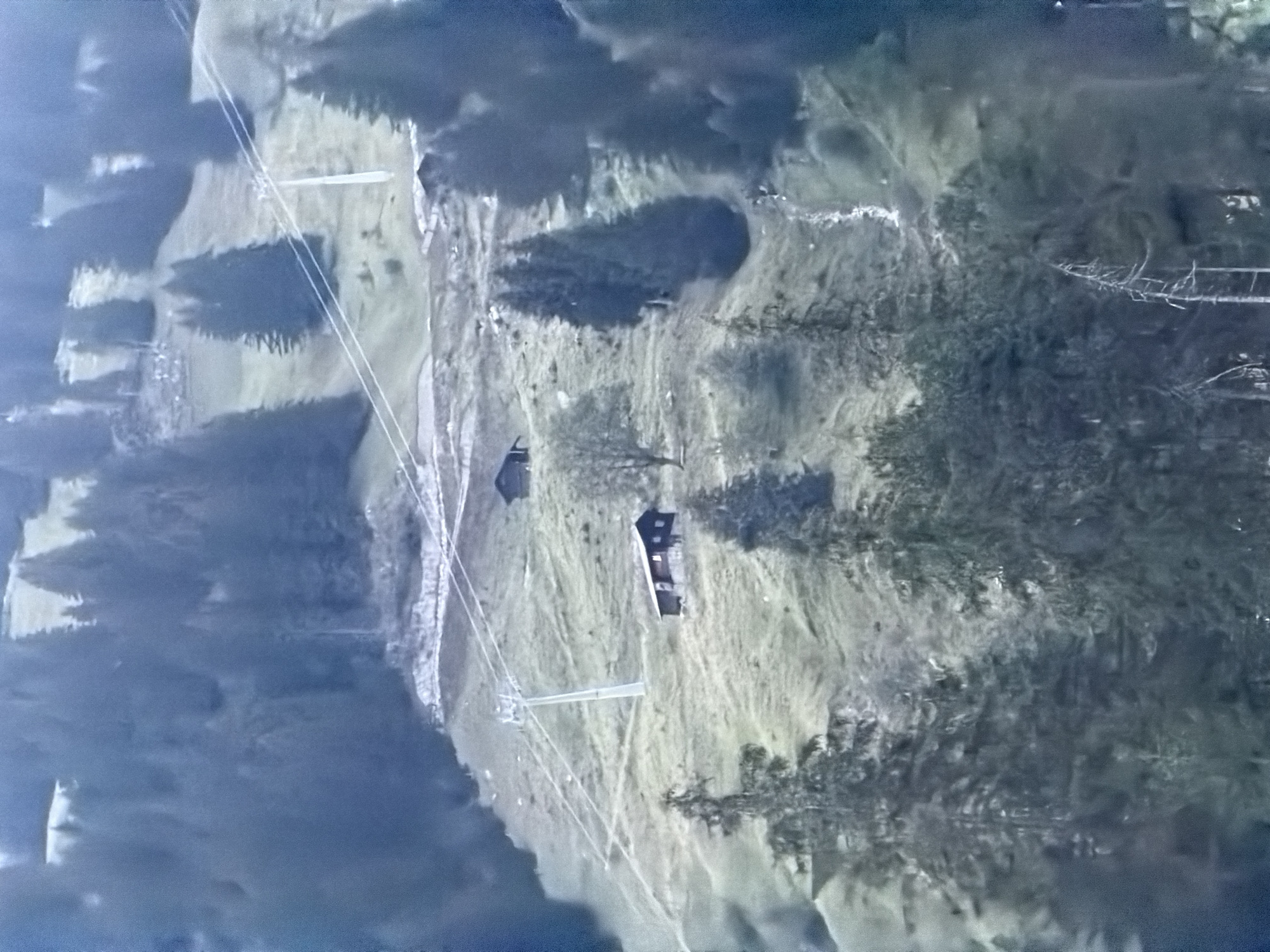 The image shows a mountainous landscape with a cable car system running up a grassy slope. Two small wooden buildings are visible on the slope, one with a dark roof and windows. Tall evergreen trees are scattered across the landscape, and a small stream runs down the hillside. The scene appears to be in a rural, mountainous area.