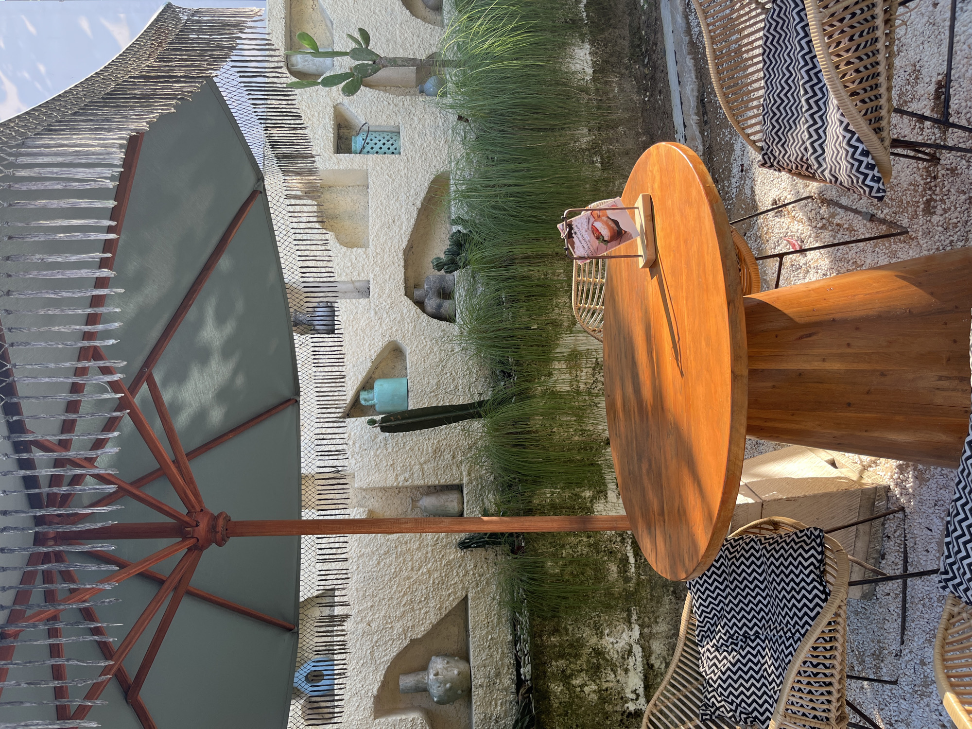 In Kuta Selatan, Indonesia, an outdoor seating area features a round wooden table with a checkered cushion on a wicker chair. The table has a small menu holder with a card displaying an image of food. Behind the table, a wall with decorative niches holds vases and plants, including cacti. A large umbrella with fringed edges provides shade. The ground is covered with small white pebbles and some dirt.