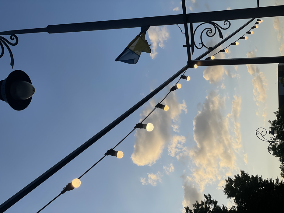 Silhouetted metal structures hold strings of lights against a blue sky dotted with white clouds. A small, faded blue and yellow flag hangs limp from one of the structures. A traditional street lamp with a wide shade is visible on the left. The lights are on, casting a warm glow. Dark green trees are partially visible at the bottom right. The perspective is looking upwards, creating a sense of openness.