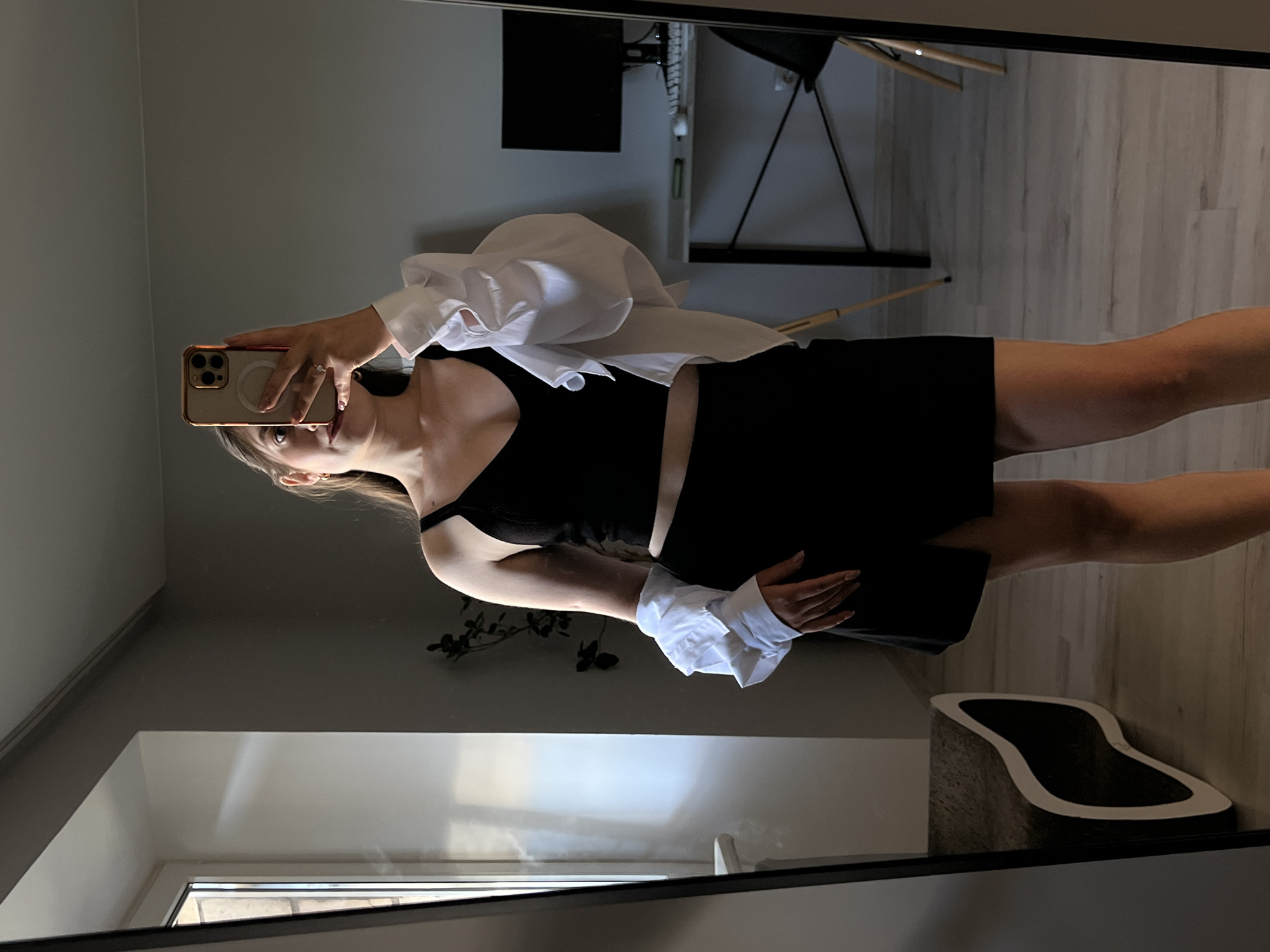 A young woman is standing in front of a mirror, taking a selfie in what appears to be a bedroom. She is wearing a black tank top and a black skirt. A white shirt is draped over her shoulders. In the background, a desk with a computer monitor and keyboard is visible, along with a chair. The room has light-colored wooden floors and grey walls. There is also a plant in the background. The woman is holding a smartphone in her right hand to take the picture. The overall impression is a casual, indoor setting. The image does not provide any specific visual cues to indicate the location is Ternopil, Ukraine.