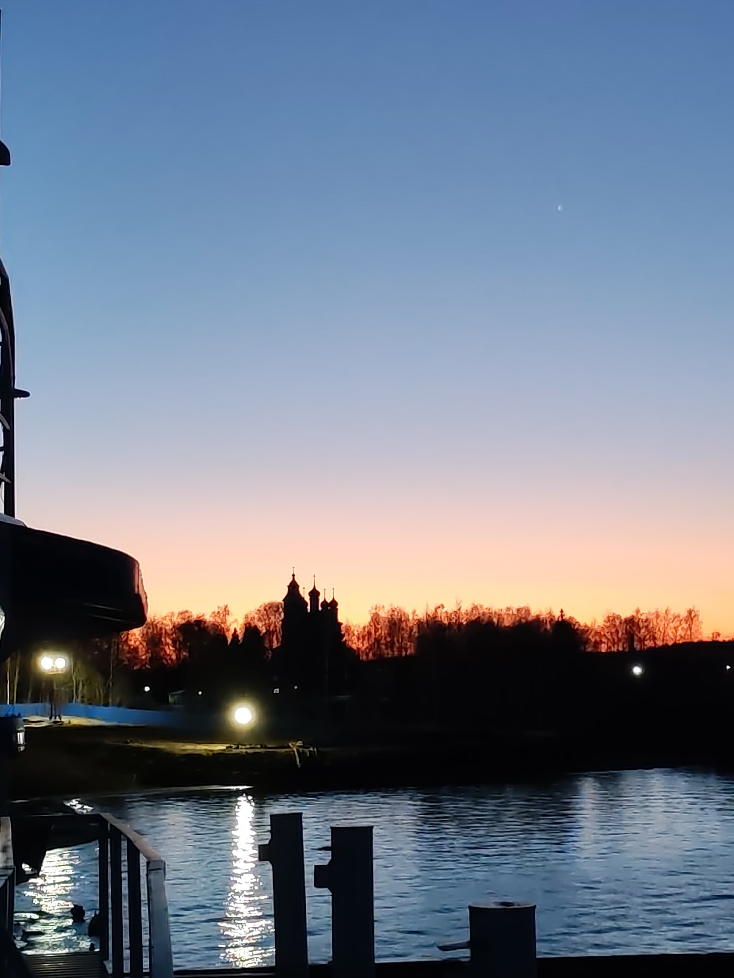 The image captures a serene twilight or dawn scene over a body of water. The sky displays a beautiful gradient, transitioning from a deep blue at the top to soft orange and pink hues near the horizon. A single, faint white dot, likely a star or planet, is visible in the upper sky.
Silhouetted against the colorful horizon are numerous bare trees and the distinctive outline of a church or monastery, characterized by its multiple domes and crosses. Below this, a calm river or canal fills the lower half of the frame, reflecting streaks of bright light from shore installations.
In the foreground on the left, a large, dark structure, appearing to be part of a boat or ship, is partially visible, with two bright circular lights illuminating its side. Mooring bollards or posts are dimly seen along the bottom edge of the image.
The distant shoreline features several artificial lights, one of which produces a strong, shimmering reflection on the water's surface. A blue tarp or structure is discernable on the left side of the distant bank, and a faint, silhouetted human figure stands near a lamp post. The scene evokes a tranquil and peaceful atmosphere. There is no visible text in the image.