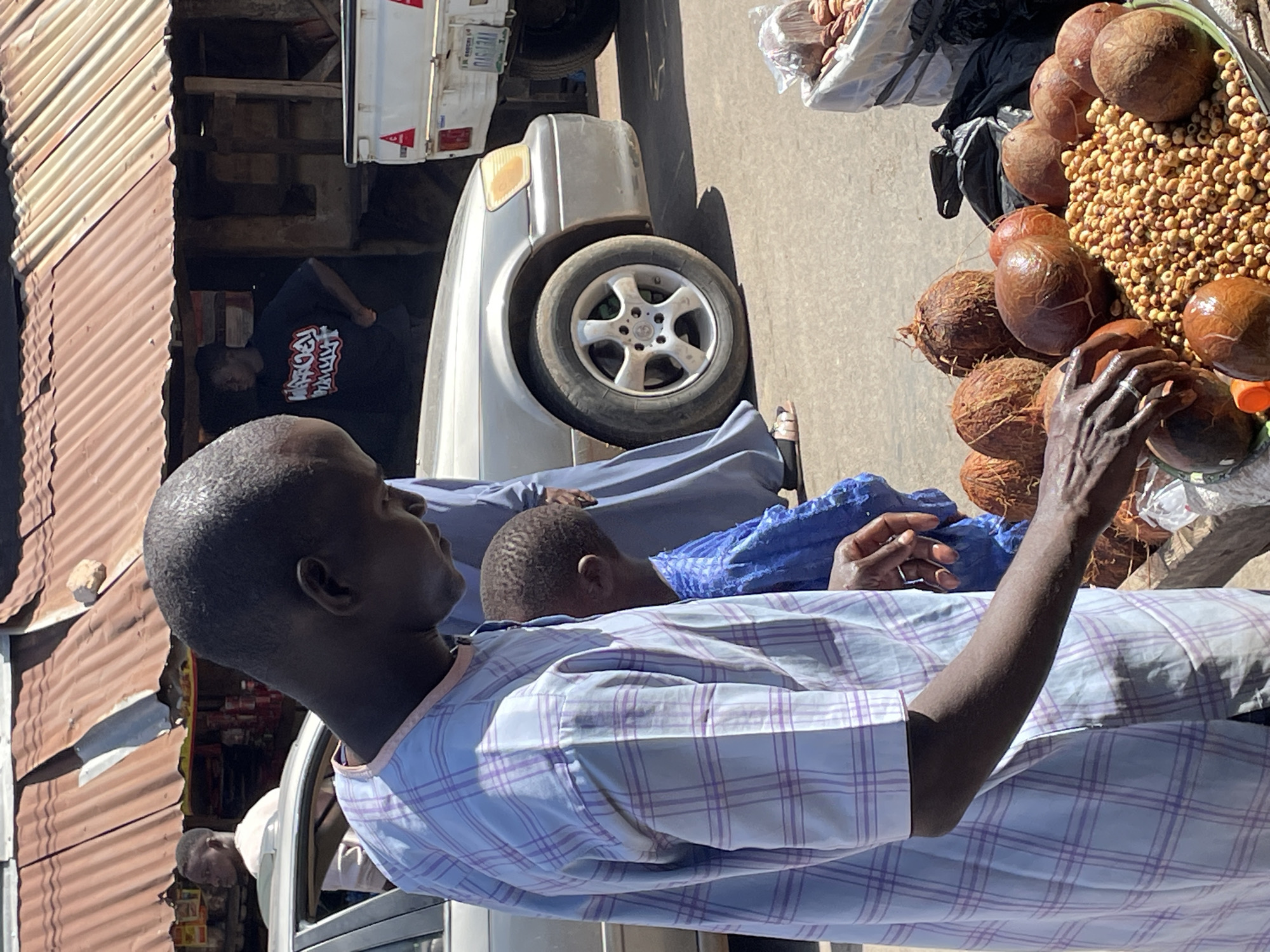 A man is seated in a white vehicle, wearing a blue and white plaid shirt, with his arm extended towards a vendor's display of kola nuts and coconuts. The vendor's produce is arranged on a table adjacent to the vehicle. The setting appears to be a street in Jos, Nigeria, with structures of corrugated metal to the left. Another person is visible in the back of the vehicle and another man is standing by the corrugated metal structure.