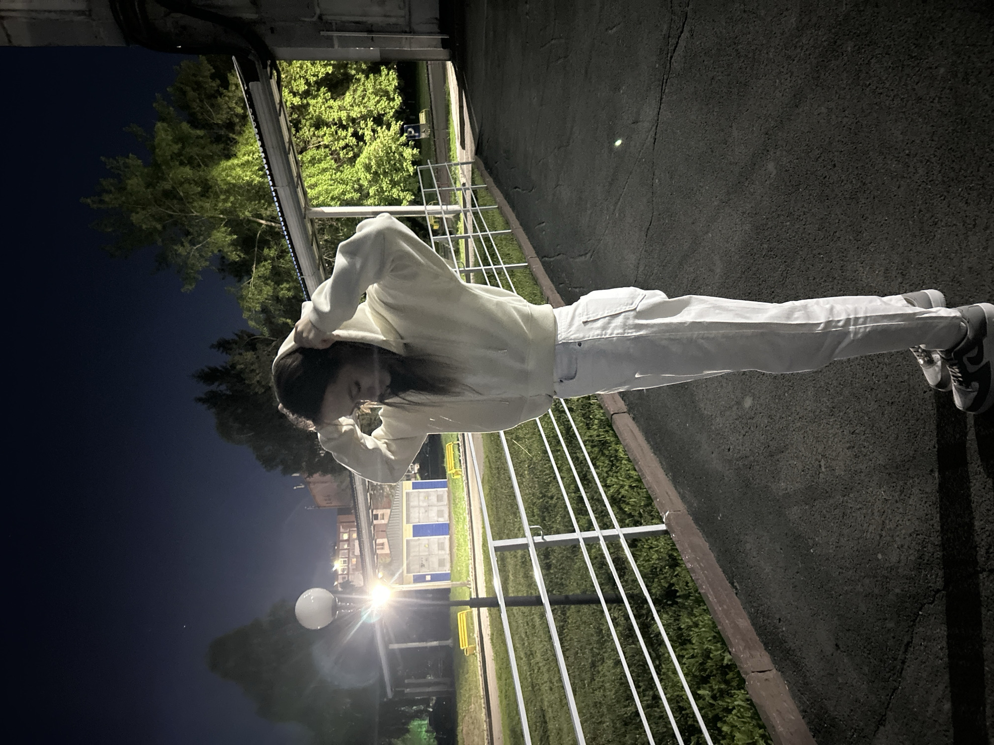 An outdoor, nighttime scene is depicted, with the image rotated 90 degrees clockwise from its natural orientation, showing a young person standing vertically on what appears to be a paved path or road. The person, positioned towards the right side of the frame, is wearing a light-colored (possibly off-white) hooded sweatshirt and matching light-colored pants. They have dark hair and are seen adjusting their hood with both hands, looking downwards, with their face partially obscured.
To the right of the person, a dark asphalt or paved surface extends, indicating the ground. To their left, a white metal fence or railing separates the paved area from a grassy expanse. Beyond the grass, trees with bright green foliage are visible, illuminated by unseen light sources. In the deeper background, especially on the left, the sky is dark blue, characteristic of night. A bright, spherical street lamp stands prominently on the mid-left side, casting a strong glow. Further back on the left, distant buildings with visible windows and some industrial-looking structures (possibly blue and yellow containers or facades) are discernible. The overall setting appears to be an urban or suburban park or pathway. The weather seems clear and calm. No visible text is present in the image.