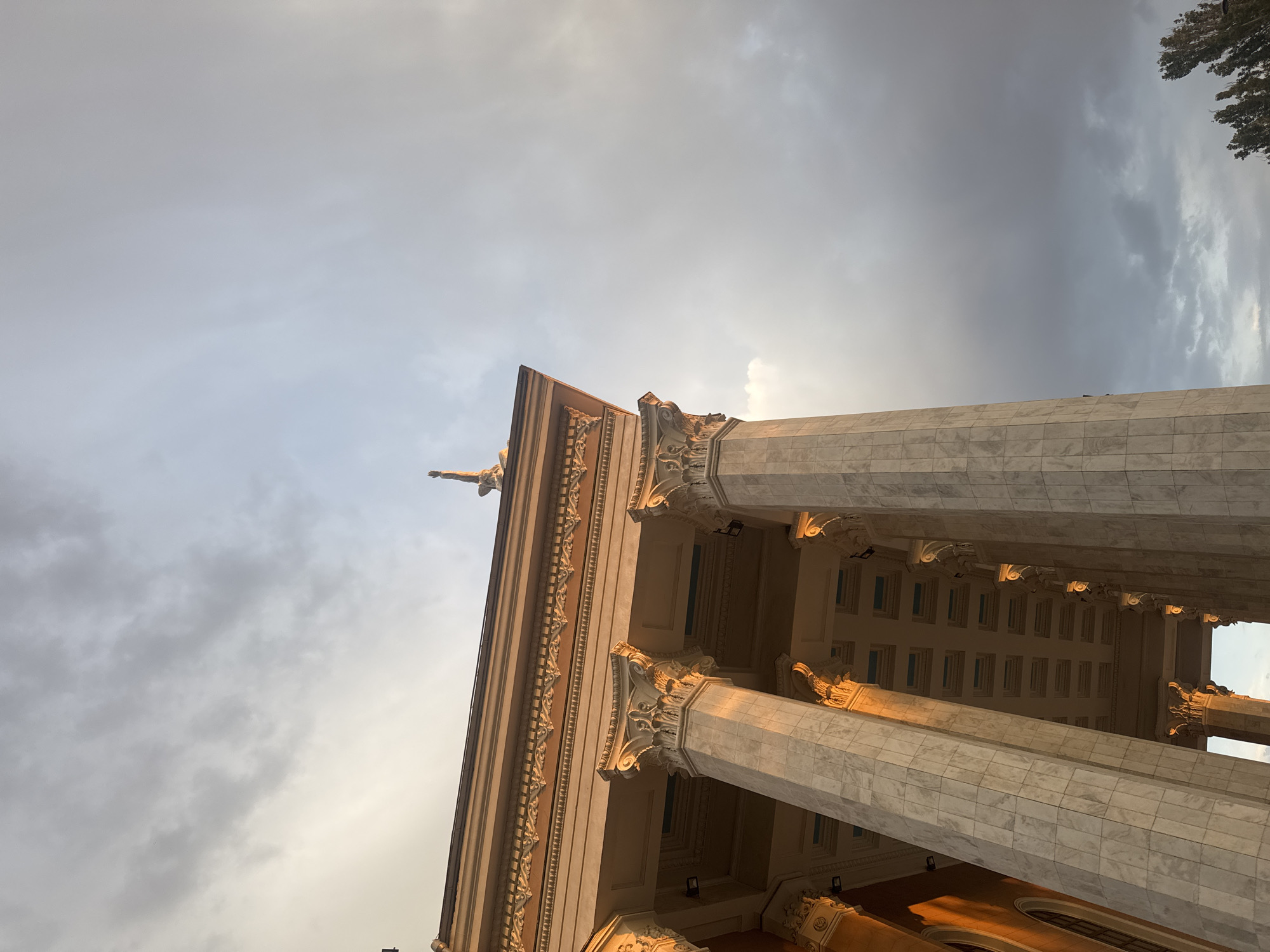 A low-angle shot captures the facade of a grand building with imposing marble columns and ornate architectural details, possibly a museum or government building, under a cloudy sky. The building's upper portion, including the roofline and decorative frieze, is illuminated by the warm glow of sunset. A slender, silhouetted statue stands on the rooftop. The sky is a gradient of light grey at the bottom, transitioning to a brighter, overcast sky with streaks of clouds overhead. To the right, the tops of a few trees are visible, their dark foliage contrasting with the muted sky. The architecture suggests a neoclassical or monumental style, common in government or cultural institutions. The overall impression is one of solemnity and grandeur, set against a dramatic sky. This building is likely the Kyrgyz State Historical Museum in Bishkek City, Kyrgyzstan, given its architectural style and prominent location.