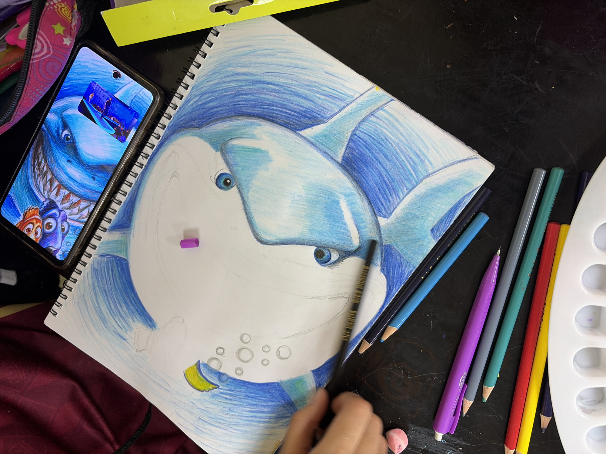 A high-angle shot depicts a work-in-progress illustration of Bruce the shark from "Finding Nemo" in a spiral-bound sketchbook. A smartphone displaying an image of Bruce alongside Nemo and Dory rests to the left of the sketchbook. The sketch predominantly utilizes blue hues. A collection of colored pencils, including shades of blue, purple, green, yellow, and red, lies adjacent to the sketchbook, along with a white paint palette. A hand holding a black colored pencil is visible in the lower right corner, implying that someone is actively drawing. The scene is set on a dark, possibly wooden surface. A portion of a pink backpack and what appears to be part of a reddish-burgundy blanket or pillow are also visible along the edges of the frame. The specific location within Guayana City, Venezuela, cannot be determined from this image.