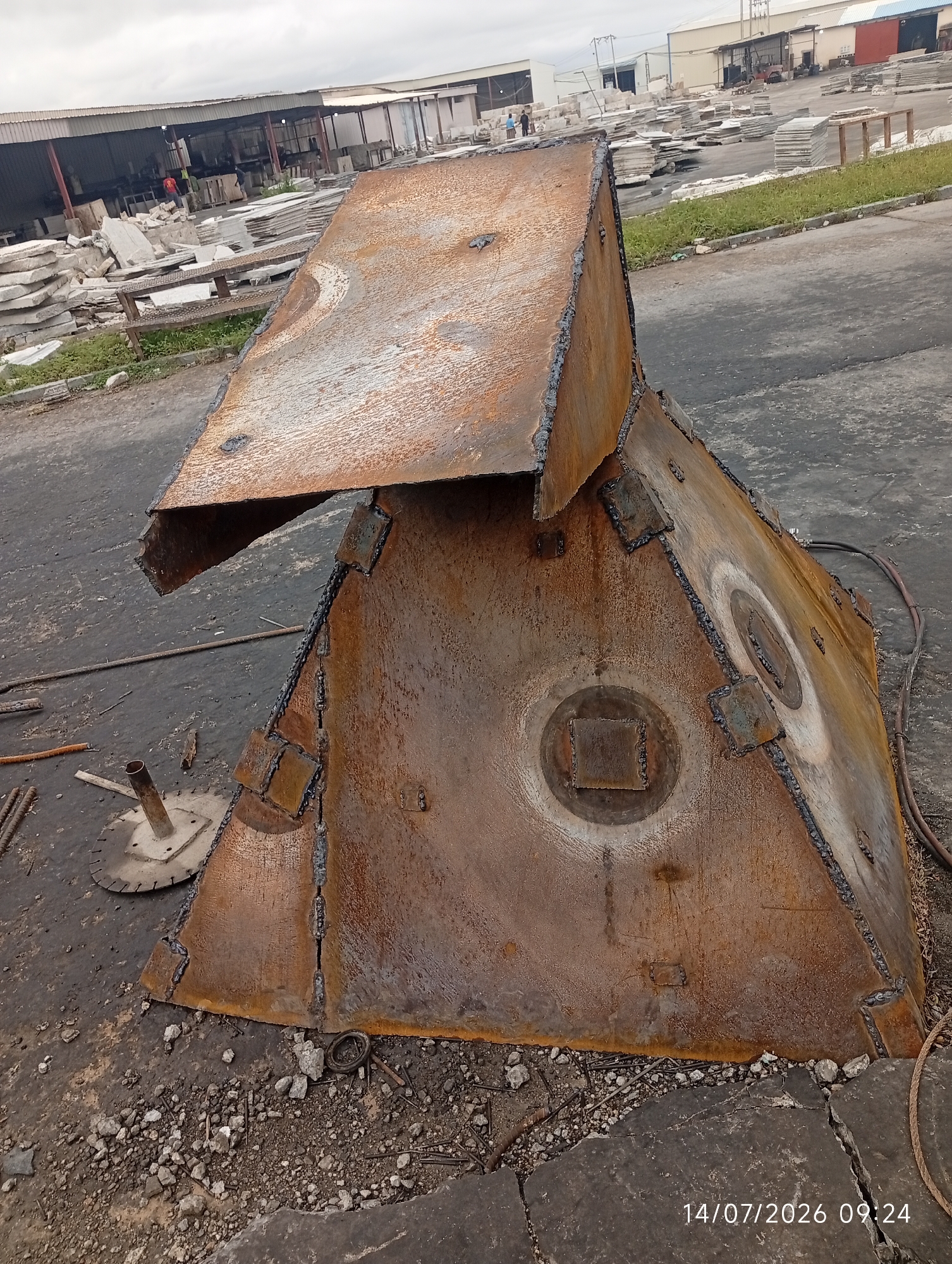 In the city of Bana Stream, Nigeria, an outdoor industrial scene unfolds. In the foreground, a large, rusty, and welded metal structure, shaped somewhat like a tent or a collapsed hopper, dominates the frame. It is composed of several joined metal plates, evident by the numerous visible welds. Two circular indentations, one with a square cutout in its center, are present on one of its large faces. The ground around the structure is a mixture of dark asphalt and gravel, littered with metal debris, including rebar and a circular saw blade with a spindle. To the left and in the background, stacks of light-colored stone slabs are arranged, suggesting a stone quarry or processing facility. In the mid-ground, industrial buildings with corrugated roofs are visible. A few individuals are sparsely present in the background, one wearing a red shirt and another in a blue shirt, appearing to be workers. The sky is overcast. The image is timestamped 14/07/2026 09:24.