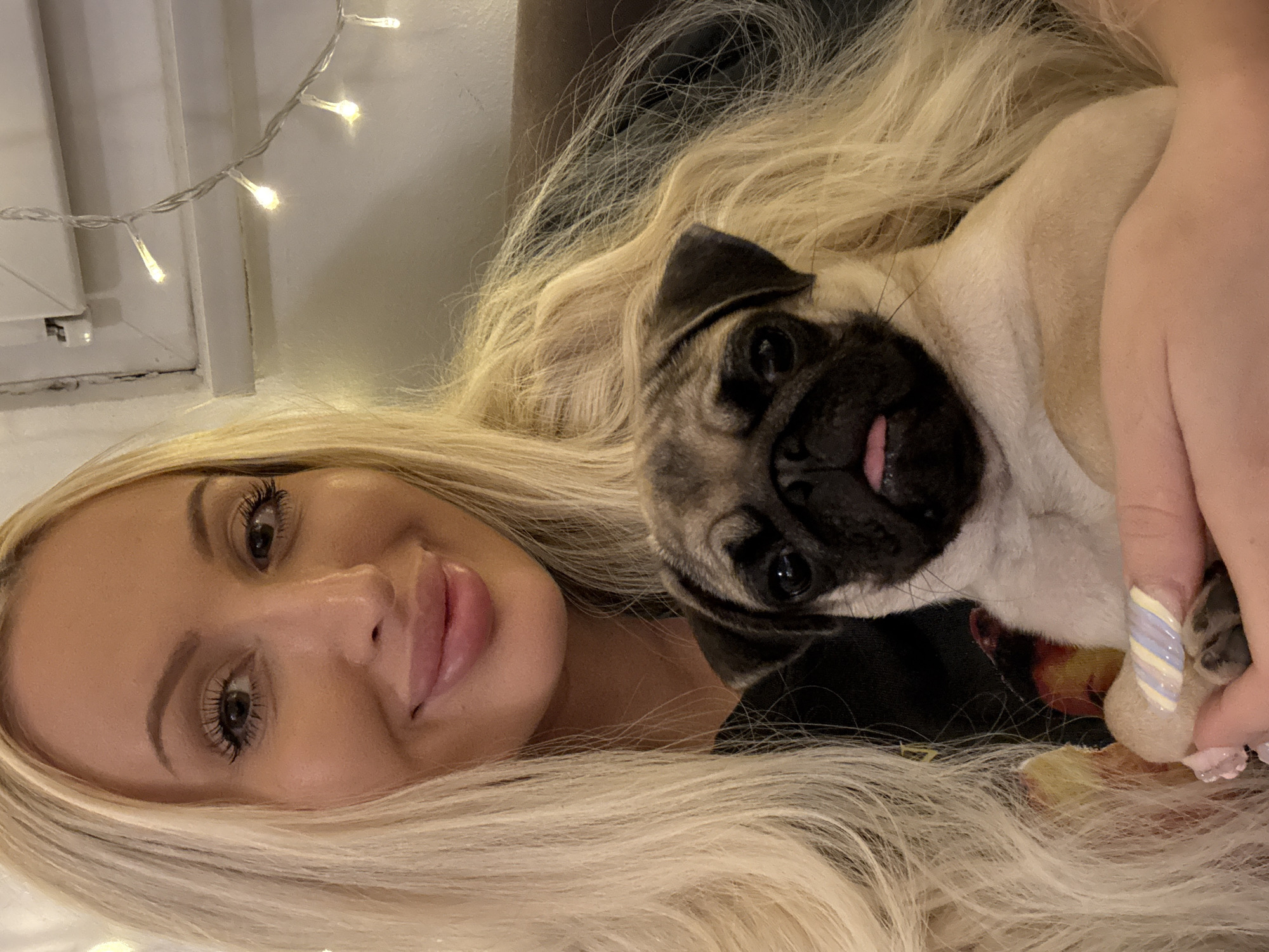 A medium shot captures a woman with long blonde hair and a pug puppy. The woman is smiling and looking at the camera, with her blonde hair cascading around her shoulders and framing the puppy. The pug puppy is held in her arms, facing the camera with its tongue sticking out slightly. The background is dimly lit, with some fairy lights visible on the right side of the frame, suggesting an indoor setting. The woman is wearing a black top with a colorful design. The puppy's paws are visible, with one paw resting on the woman's hand. The scene appears to be a casual and affectionate moment between the woman and her pet.