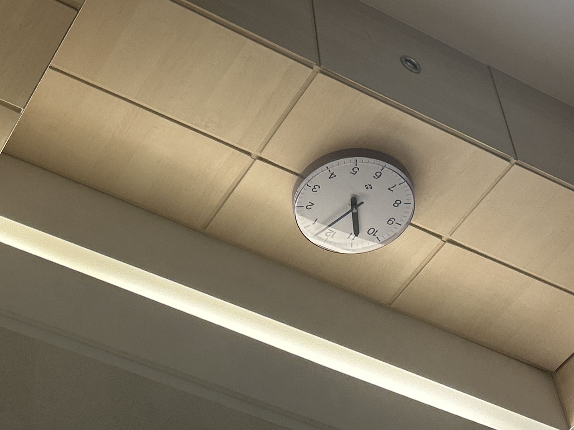 The image shows a wall clock on a wood-paneled wall. The clock is round with a white face and black hands. The time is approximately 10:48. The wall is angled, and there is a bright light source on the left side of the image, which appears to be a fluorescent light fixture. The setting is indoors, possibly an office or a public building. No specific location in Gummersbach, Germany, can be identified from the image.