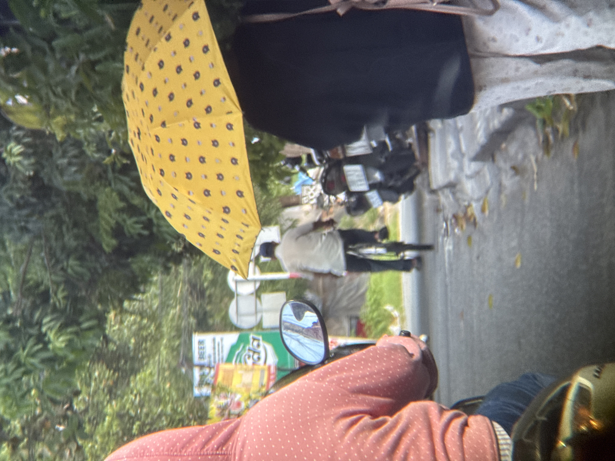 The image depicts a street scene in An Cựu Ward, Vietnam. In the foreground, a person wearing a pink hooded jacket with white polka dots is seated on a motorcycle, viewed from behind. The motorcycle's side mirror reflects a person in a white shirt. Ahead, a person in a light-colored shirt and dark pants is riding a bicycle. This cyclist is partially obscured by a yellow umbrella adorned with small dark patterns, held by someone standing to the right of the frame, wearing a dark garment and patterned pants. In the background, lush green foliage forms a dense canopy, and a sign that appears to advertise "BEER" is visible. Several motorcycles are parked on the side of the road. The ground is paved and appears damp in places, with fallen leaves scattered on the surface. The lighting suggests it is daytime, possibly overcast or after rain.