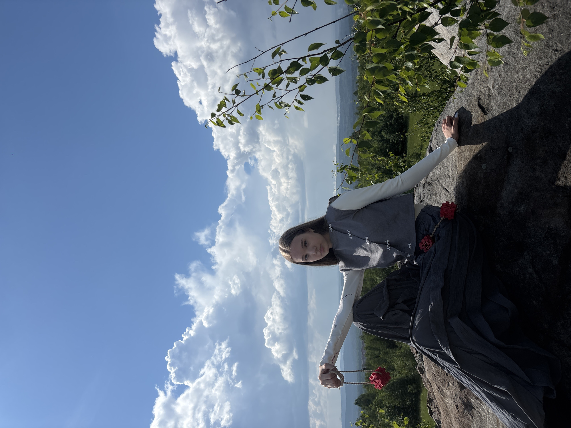 In Gubakha, Russia, a young woman with long brown hair sits on a large rock formation. She is wearing a white long-sleeved shirt under a gray sleeveless vest with decorative clasps, and a dark gray, flowing skirt. In her left hand, she holds a red cubic object suspended by a rope. Another similar red object is attached to her skirt on the right side. The background features a vast expanse of blue sky with dramatic white clouds. Below the sky, a dense green forest stretches across rolling hills, with distant haze indicating more landscape beyond. The lighting suggests it is daytime with bright sunshine casting shadows.