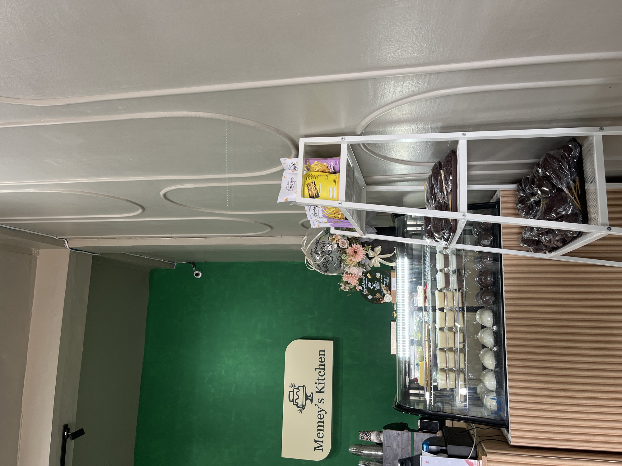 The image shows the interior of a bakery or cafe, likely located in Lhokseumawe, Indonesia. A prominent sign on the green wall reads "Memey's Kitchen". In the foreground, a refrigerated display case is filled with various desserts, including what appear to be cakes, puddings, and individual pastries. To the right of the display case is a white, multi-tiered shelving unit. The shelves hold packaged snacks and larger, individually wrapped food items, possibly baked goods like cakes on sticks. The walls are painted in two distinct colors: a vibrant green on the left and a textured, light gray on the right, with decorative grooved paneling. In the upper left corner, a black spotlight is visible on the ceiling. A small security camera is mounted on the green wall. No people are present in the image.