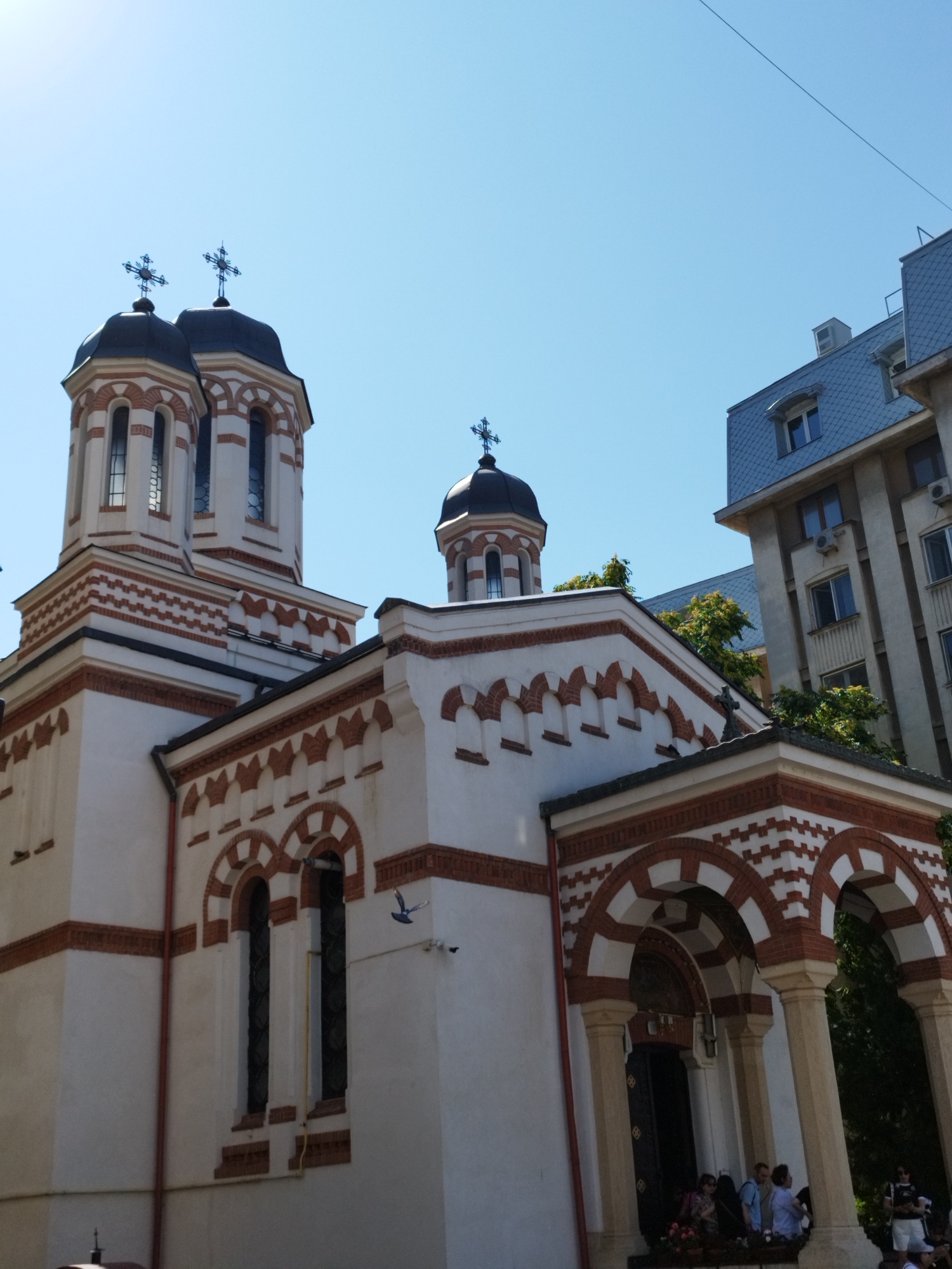 The image captures the exterior of a church in Bucharest, Romania, with a clear blue sky overhead. The church is predominantly white with red brick detailing. It features multiple domes topped with crosses, arched windows, and a portico entrance with columns. To the right of the church, a modern building with a grey, tiled roof and multiple windows is visible. In the foreground, several people are gathered near the church entrance, and a pigeon is in flight. The scene is bathed in sunlight, suggesting it is daytime.