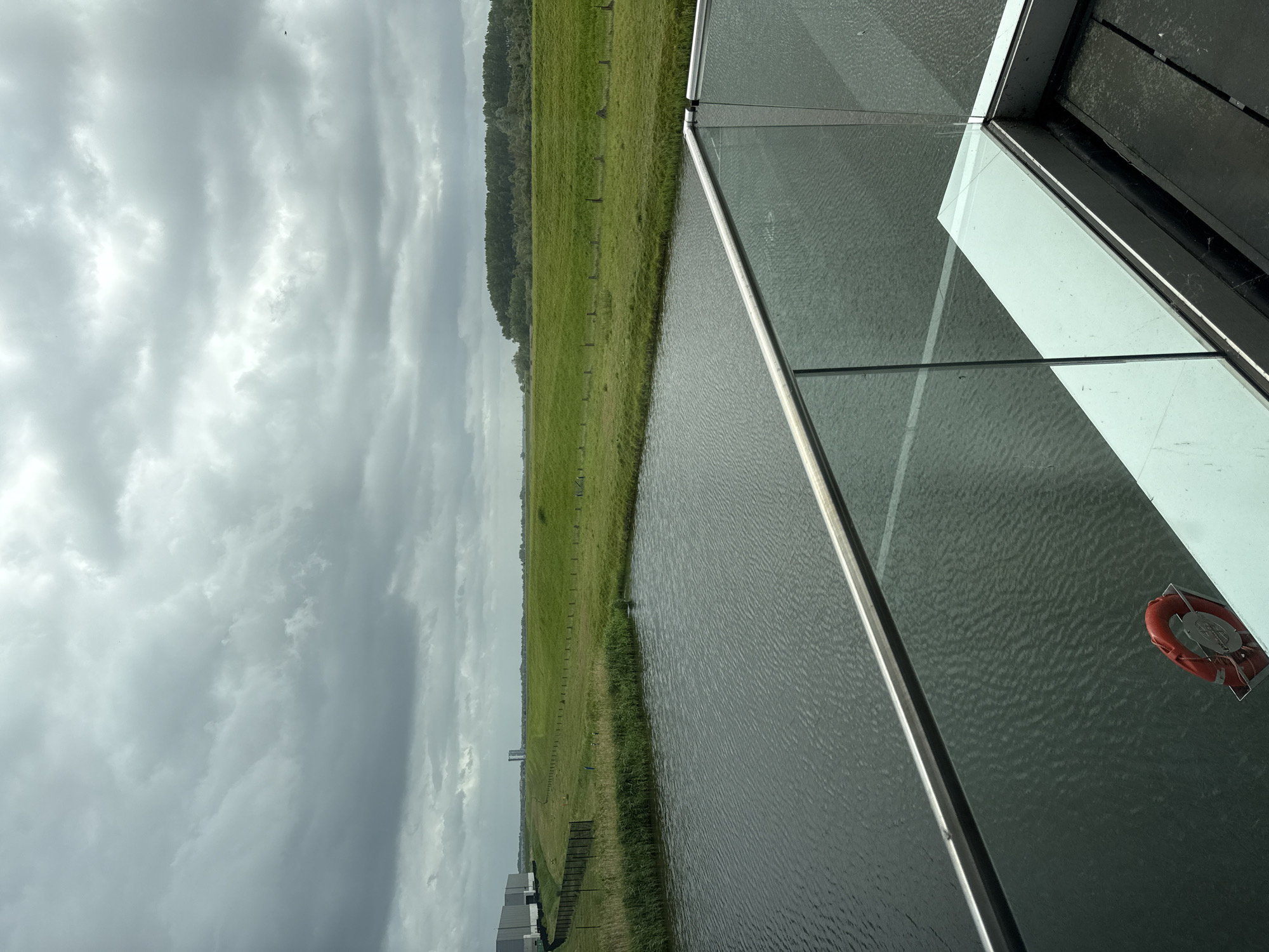 The image captures a view of a body of water, likely a canal or wide river, with green grassy banks extending into the distance. A line of trees is visible along the horizon. Above, the sky is overcast with heavy, dark clouds.
In the foreground, there is a glass barrier or balcony, possibly a part of a building or structure. A red life preserver ring is mounted on the glass panel. The location is in the city of Unknown, Unknown.